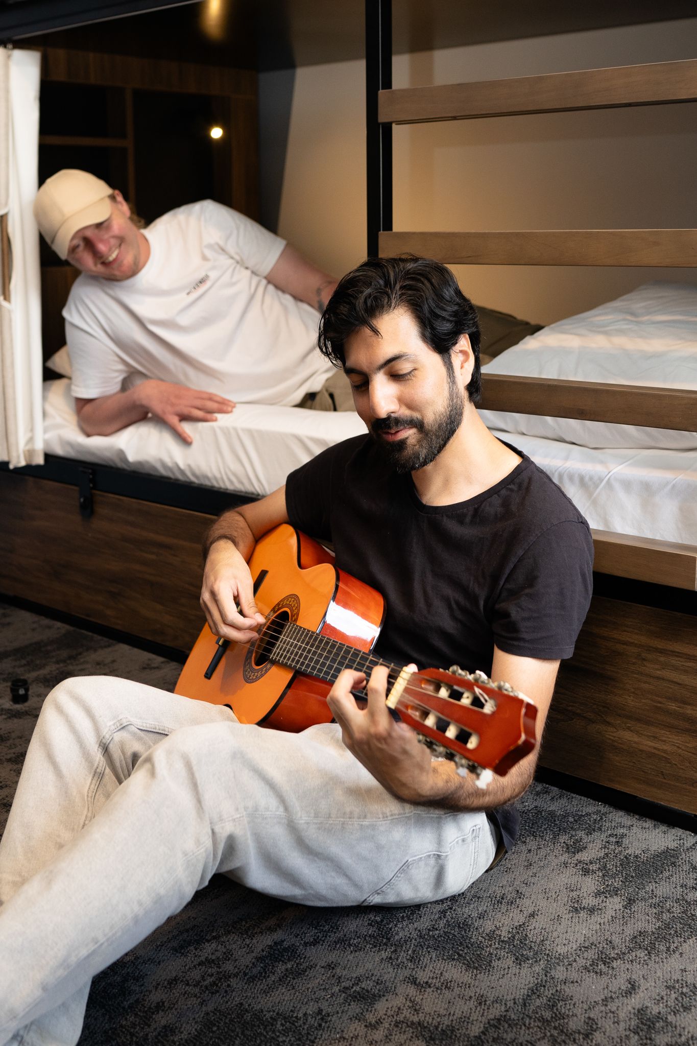 Young man playing acoustic guitar on hostel floor while friend listens from bunk bed.