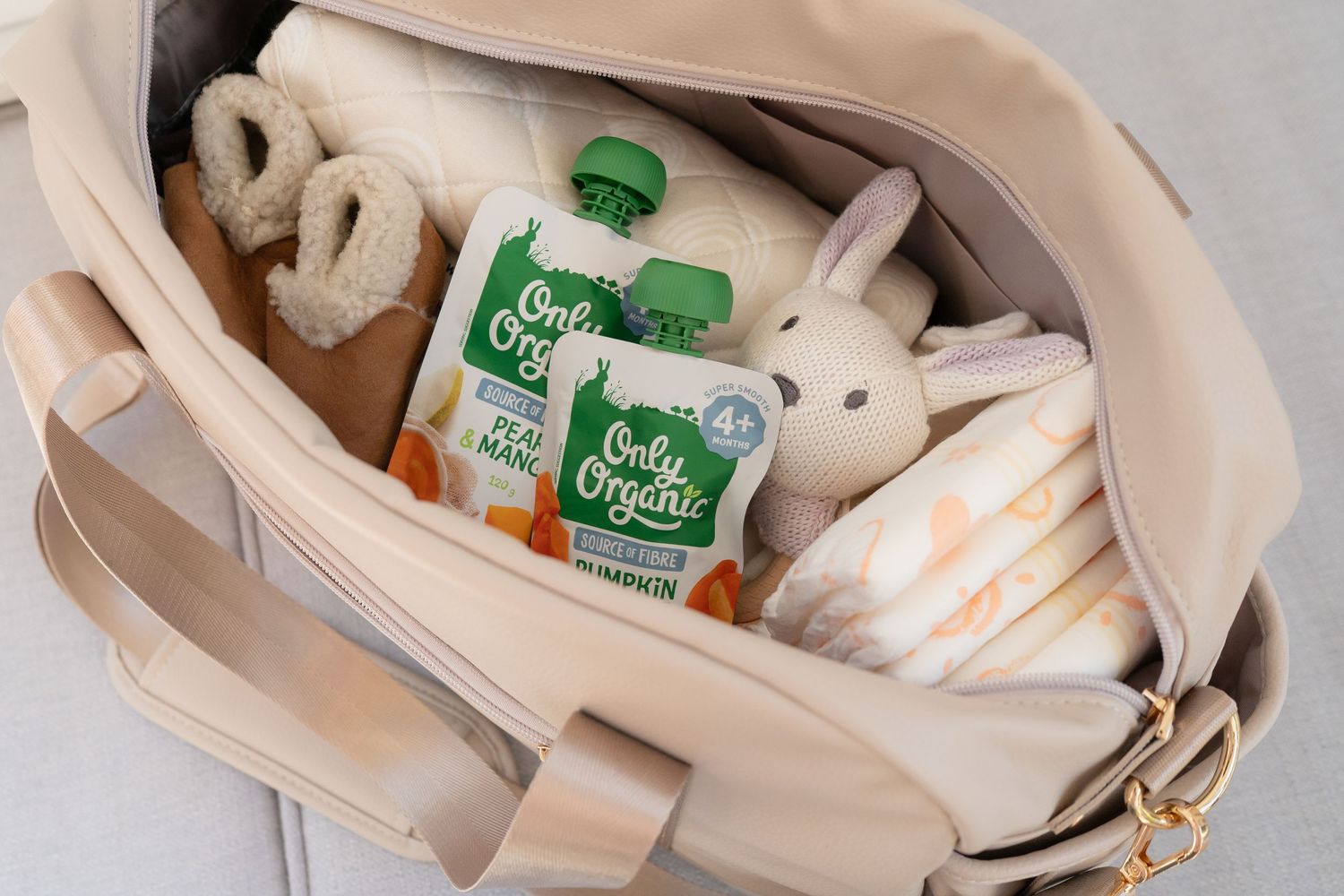 Beige diaper bag filled with Only Organic baby food pouches, teething toys, a plush bunny, and a folded blanket.