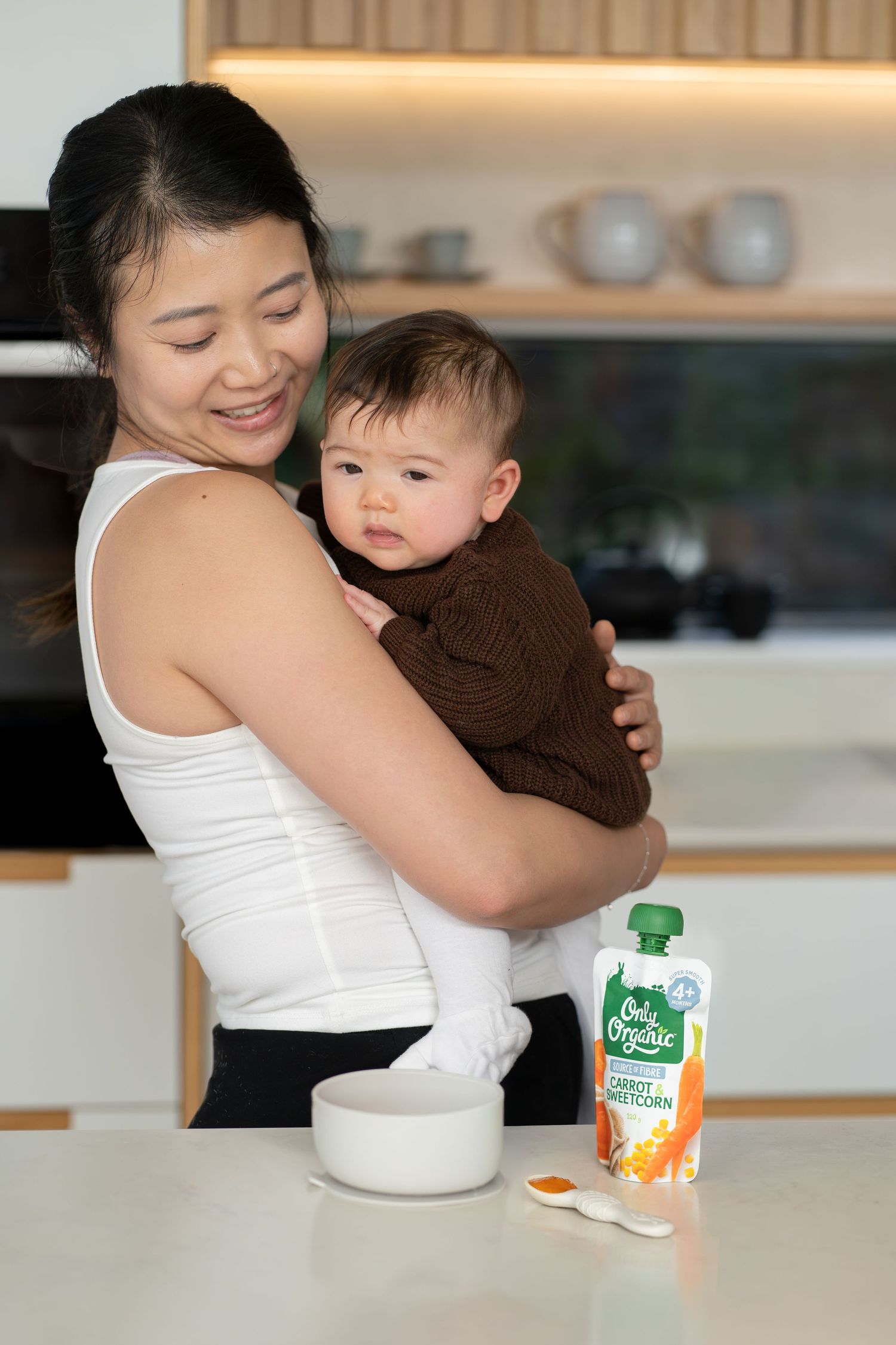 Mother holding baby in kitchen with Hello Organic carrot sweetcorn baby food pouch on counter.