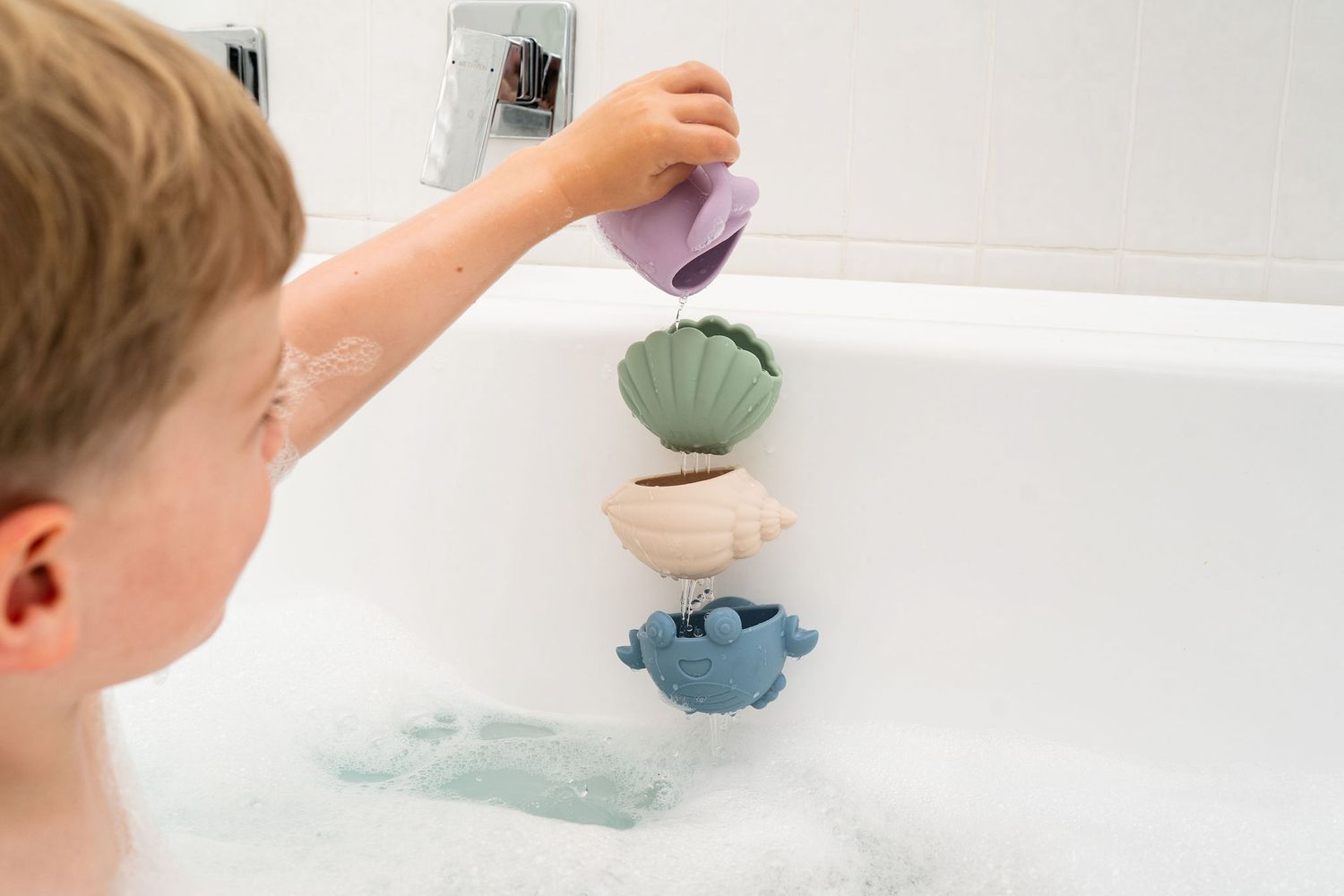 Child playing with shell-shaped stacking cups in bubble bath.