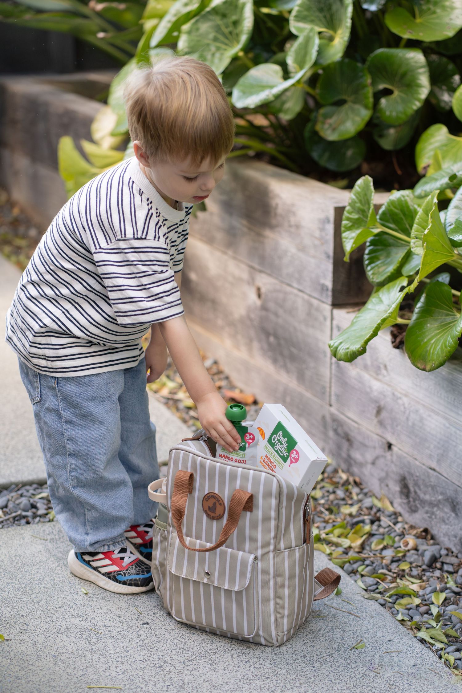 Toddler boy in striped shirt reaching into a beige striped backpack on a garden path near green plants.