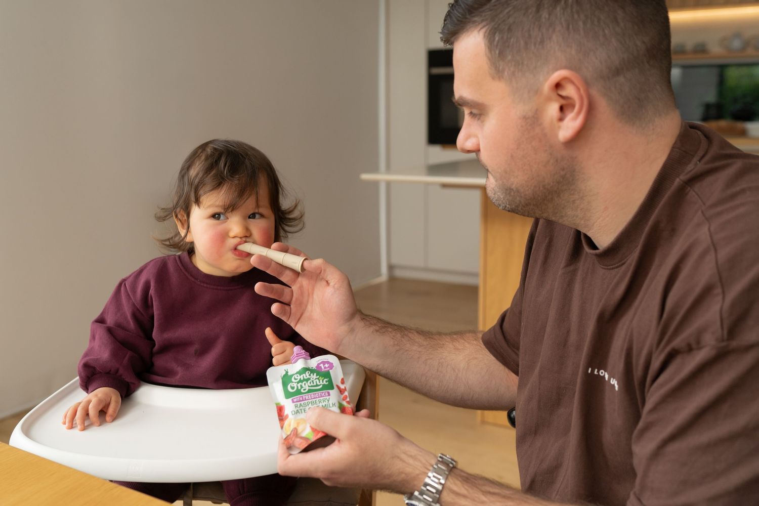Father feeding toddler in high chair with a spoon from a baby food pouch in a modern kitchen.
