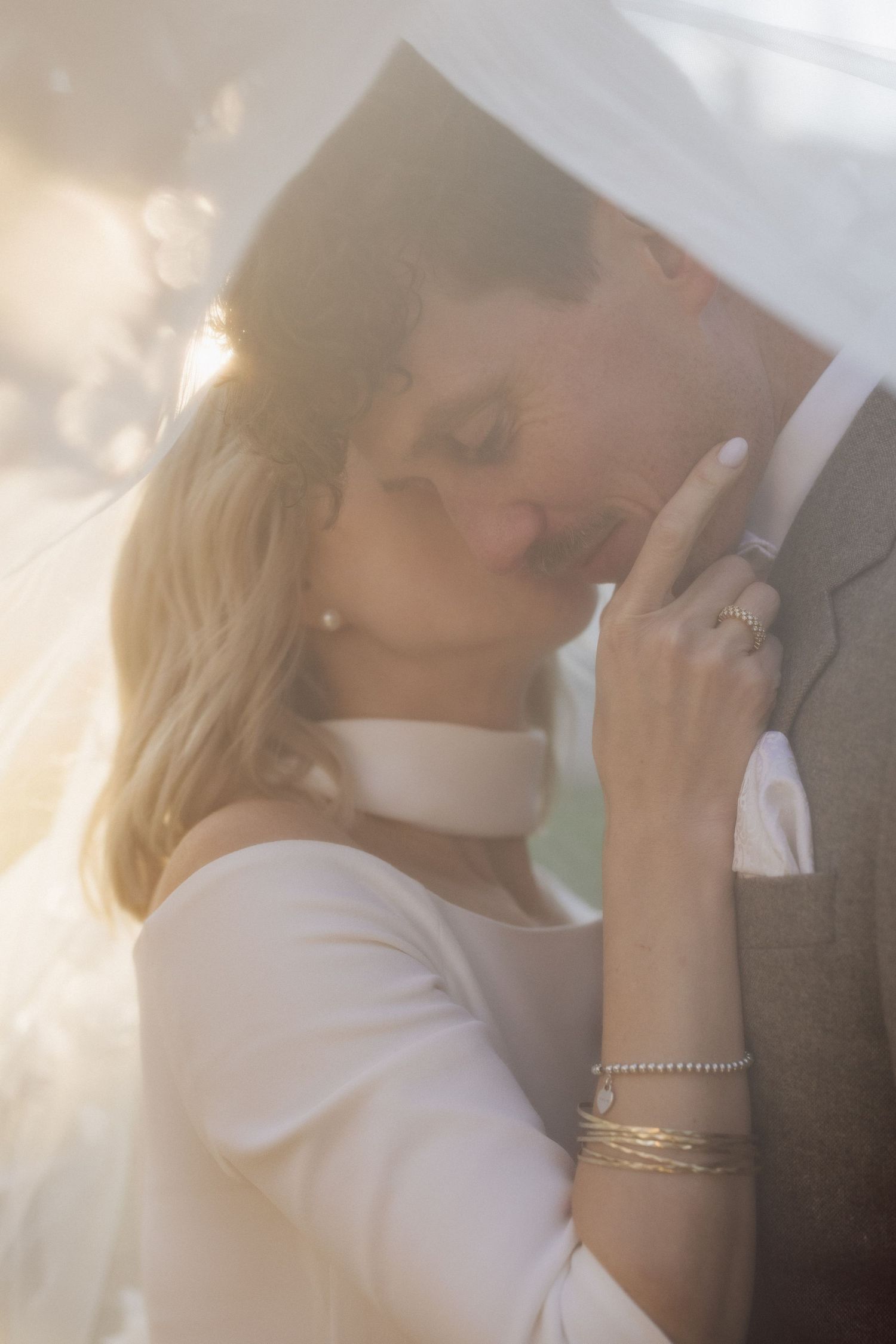 Intimate close-up of a bride and groom sharing a tender kiss under a bridal veil in warm sunlight.