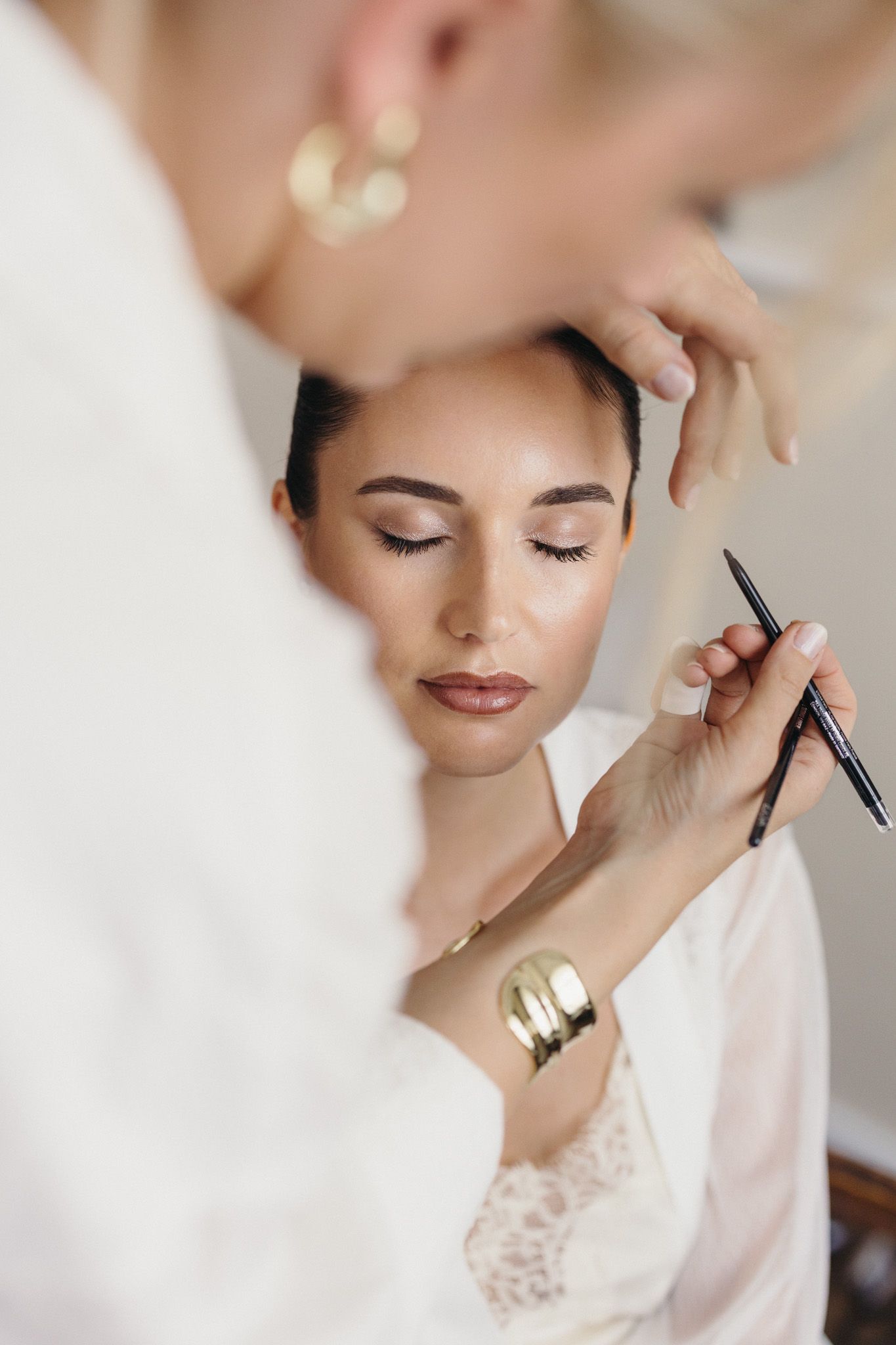 Professional makeup artist applying eyeliner to a bride in a white robe during wedding preparation.