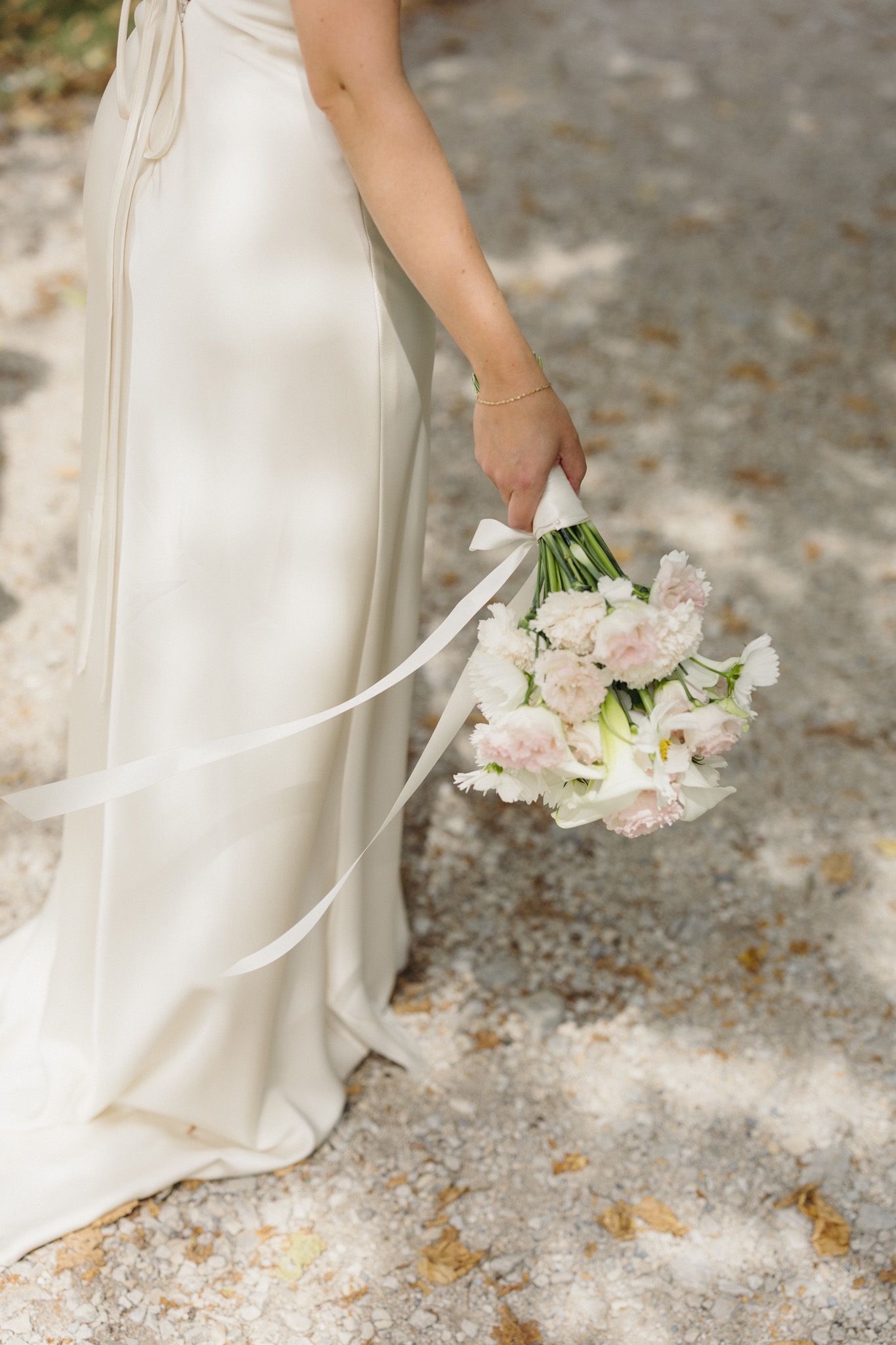 Bride in ivory gown holding delicate blush and white floral bouquet with satin ribbon on gravel path.