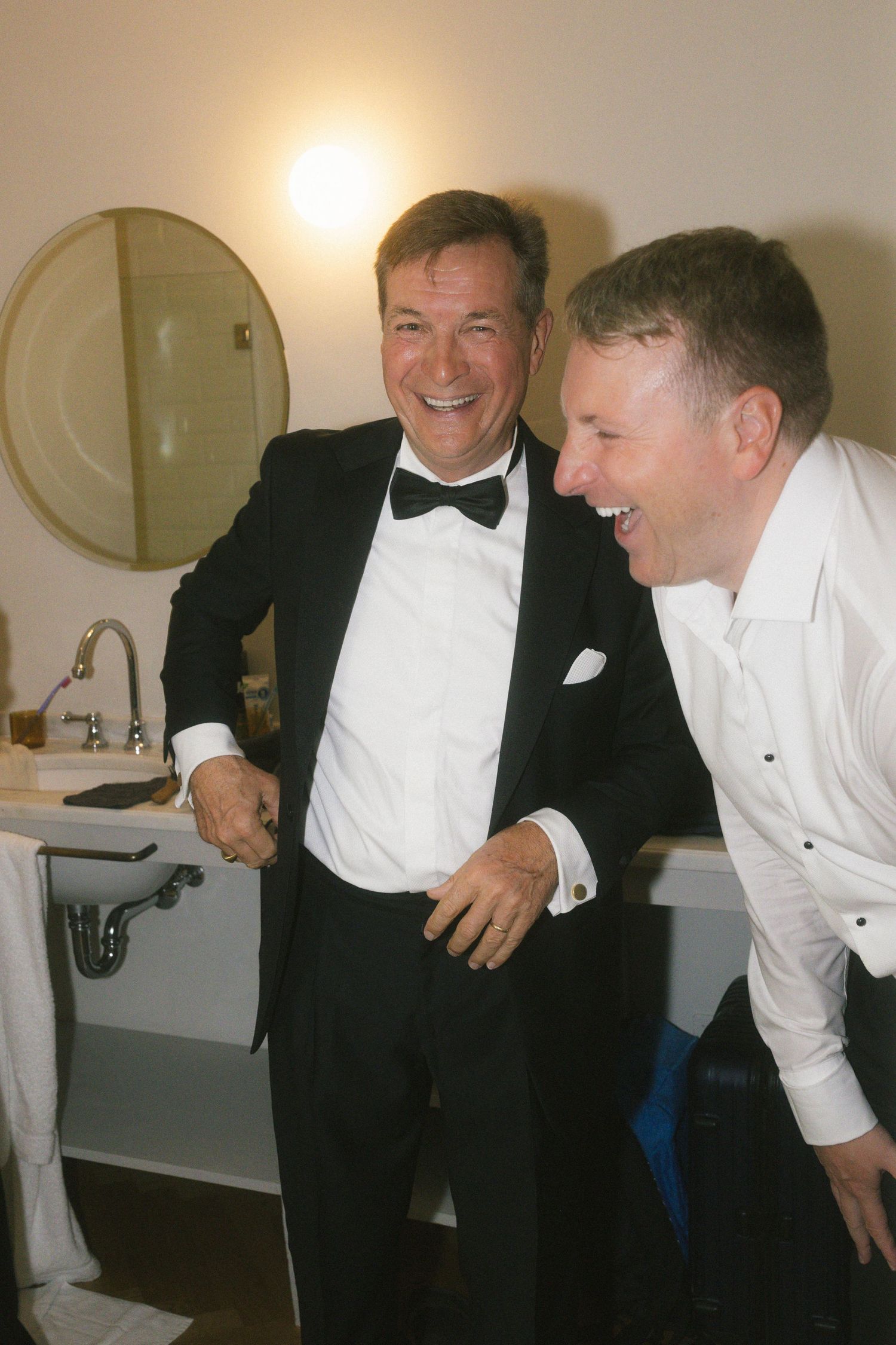 Two men laughing together in a bathroom, one dressed in a black tuxedo with bow tie, during wedding preparations.
