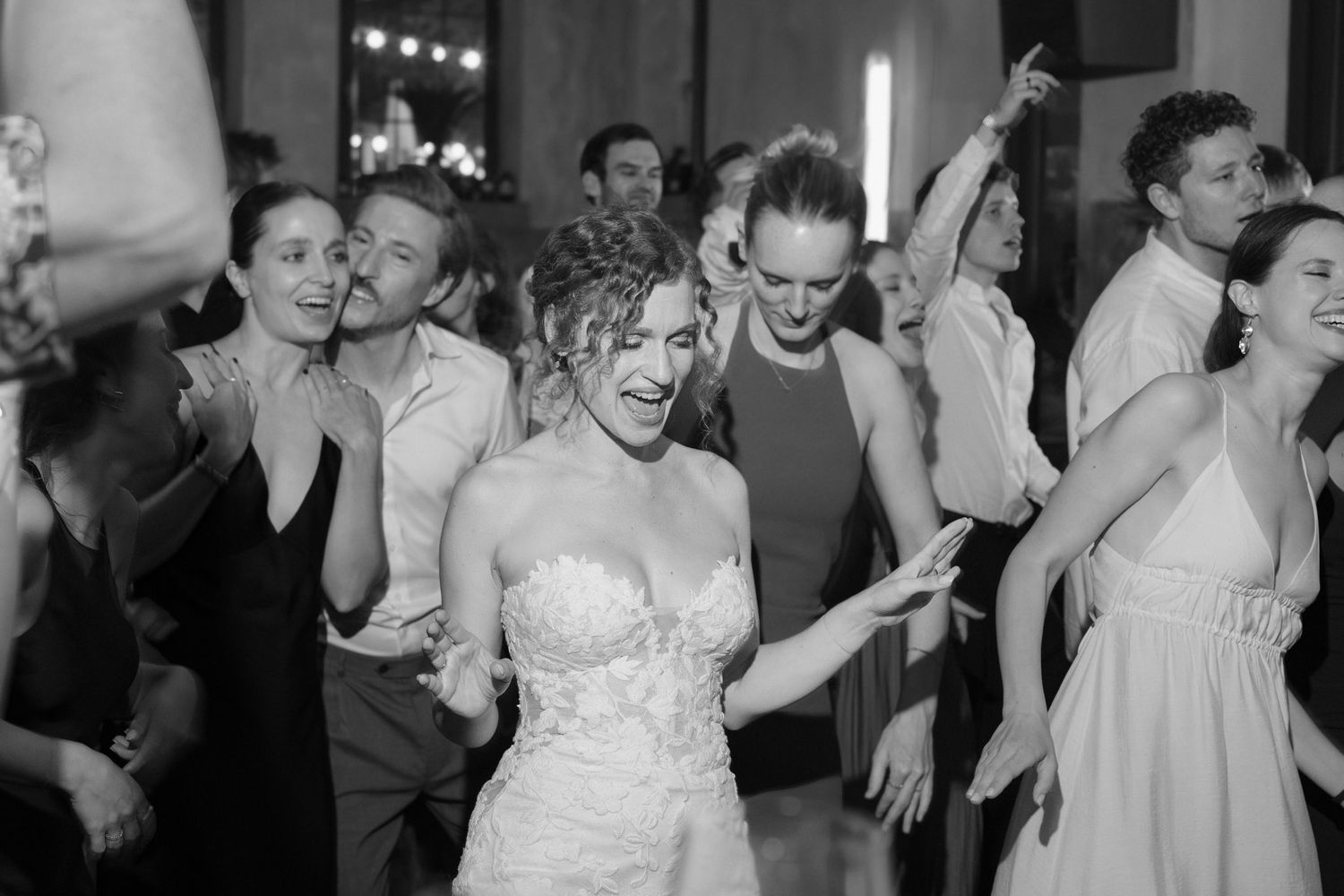 Bride in strapless lace gown dancing joyfully with wedding guests at lively black and white reception.