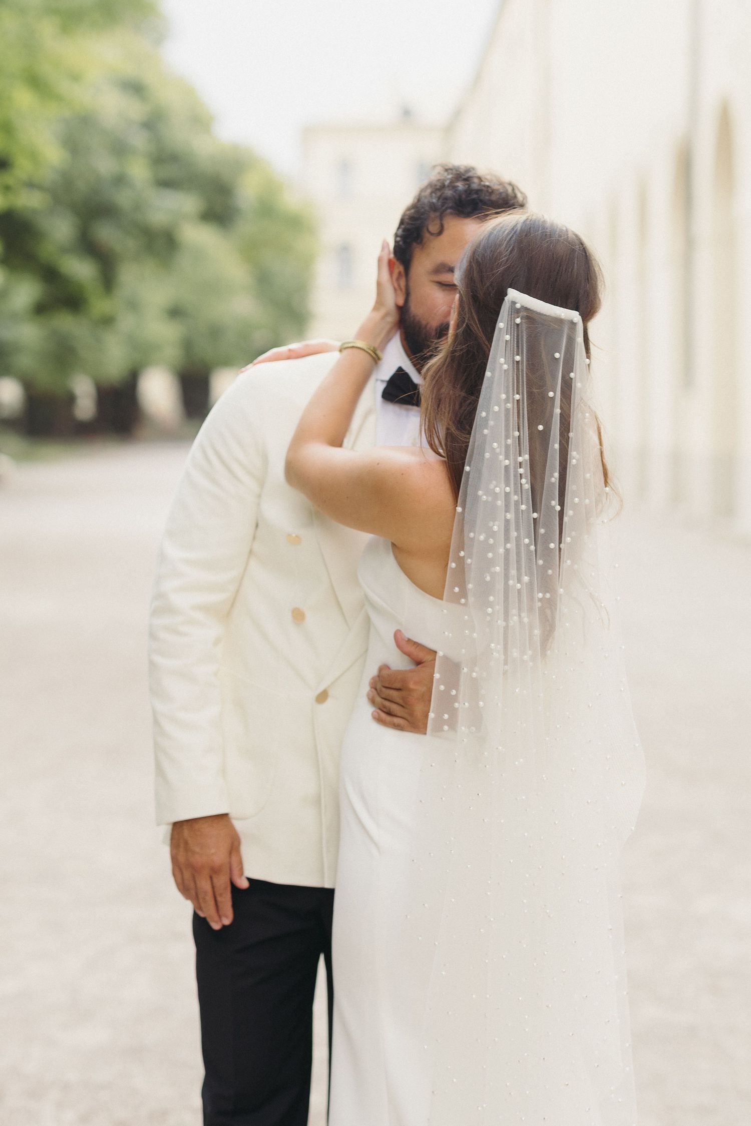 Groom in white double-breasted jacket kissing bride wearing pearl-embellished wedding veil.