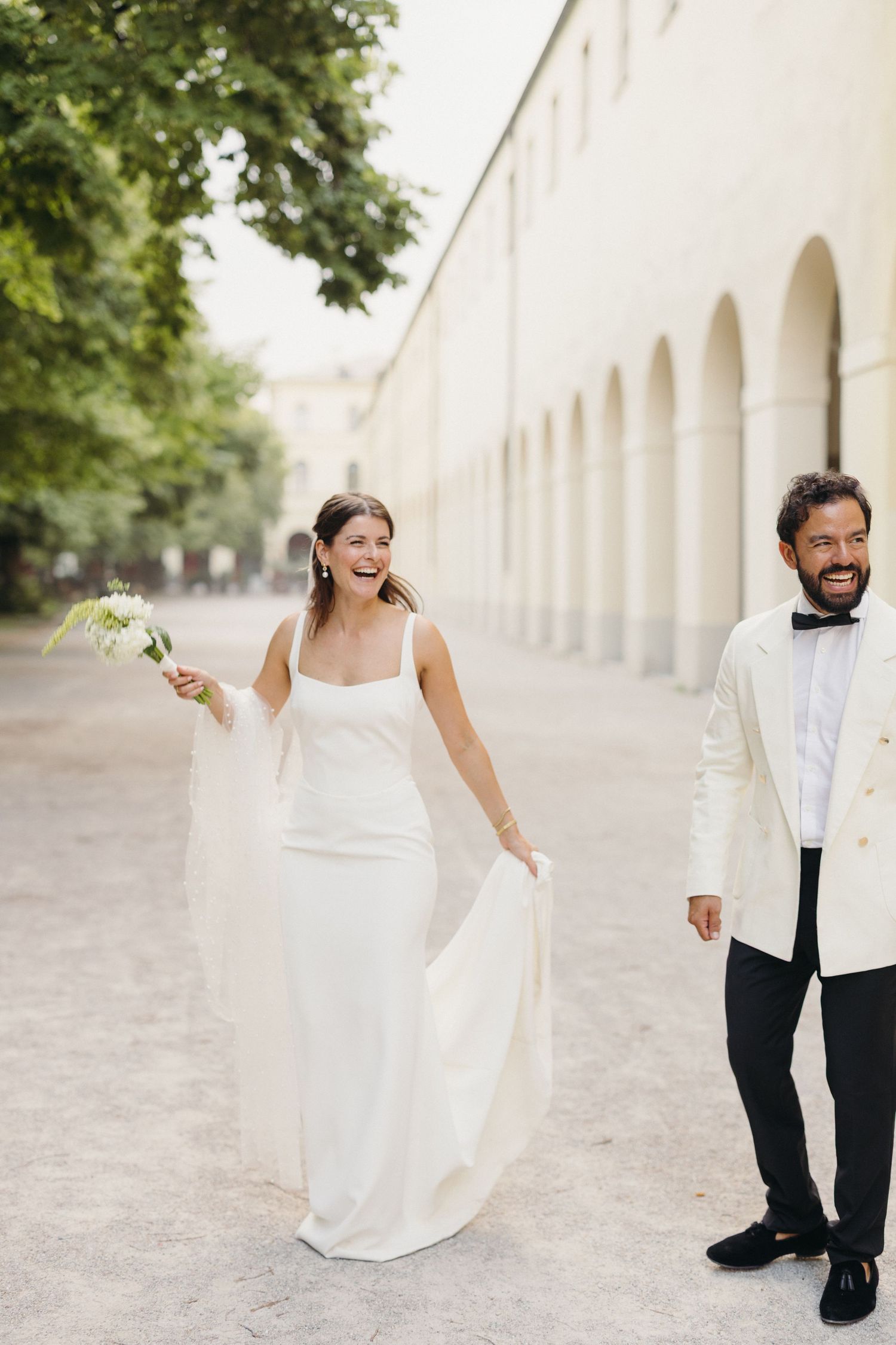 Joyful bride in white dress and groom in white tuxedo walking together past arched building exterior.