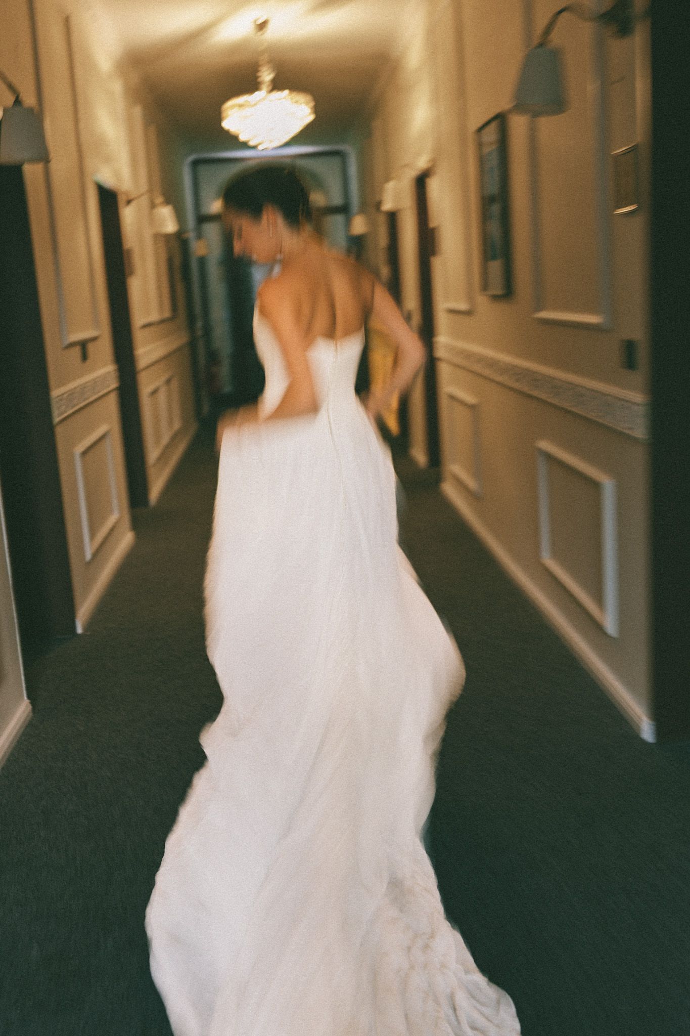 A bride in a strapless white wedding gown walks down an elegant hotel corridor, her long train trailing behind her.