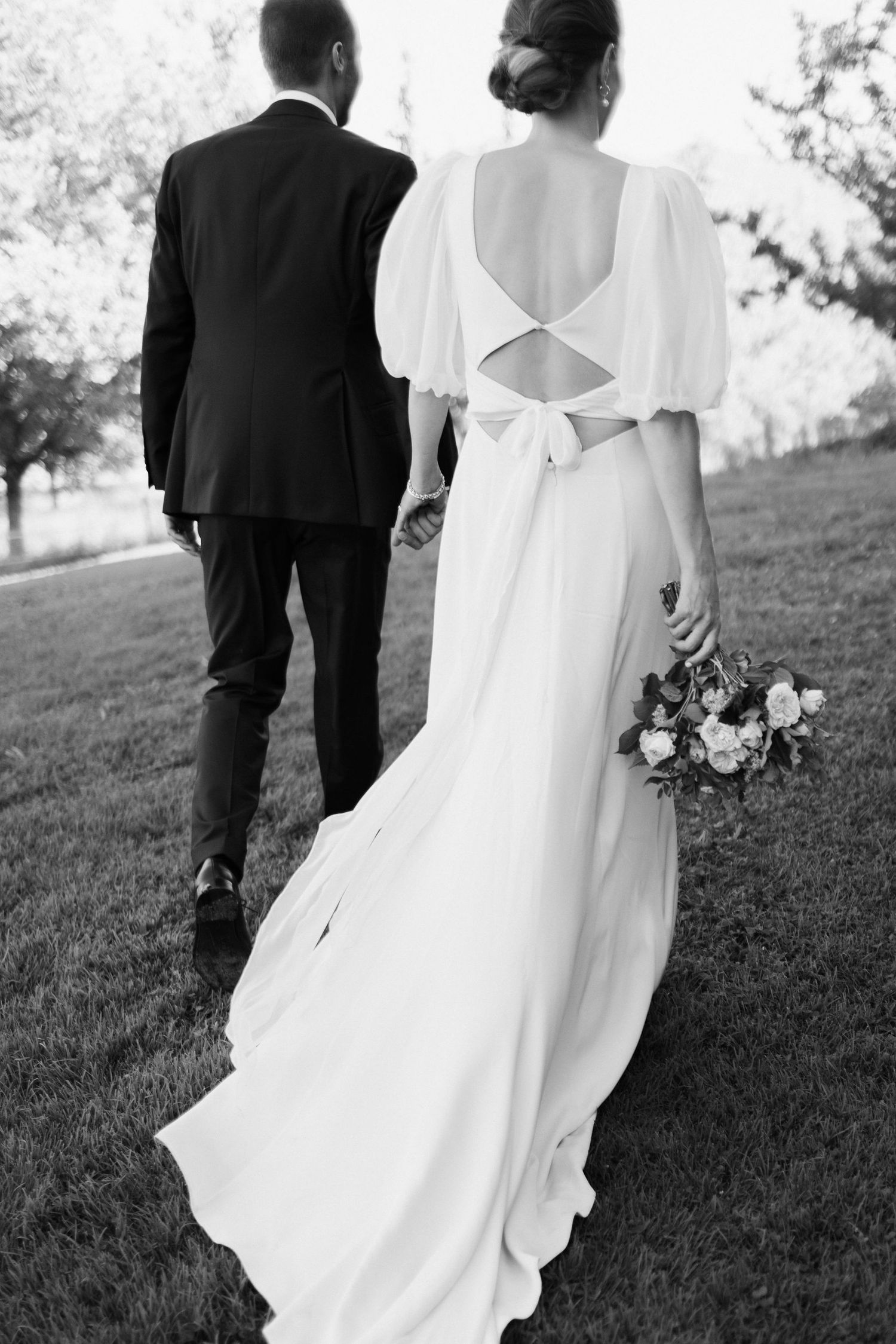 Black and white photo of bride in open-back gown holding bouquet, walking hand in hand with groom on grassy hill.