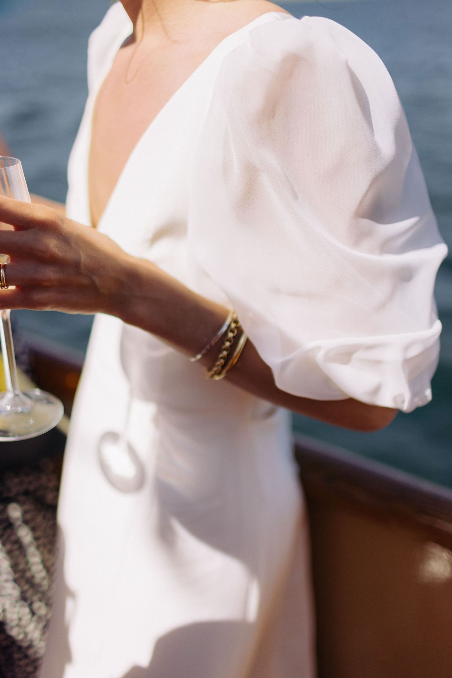 Woman in white V-neck dress with gold bracelets holding a champagne glass near water.