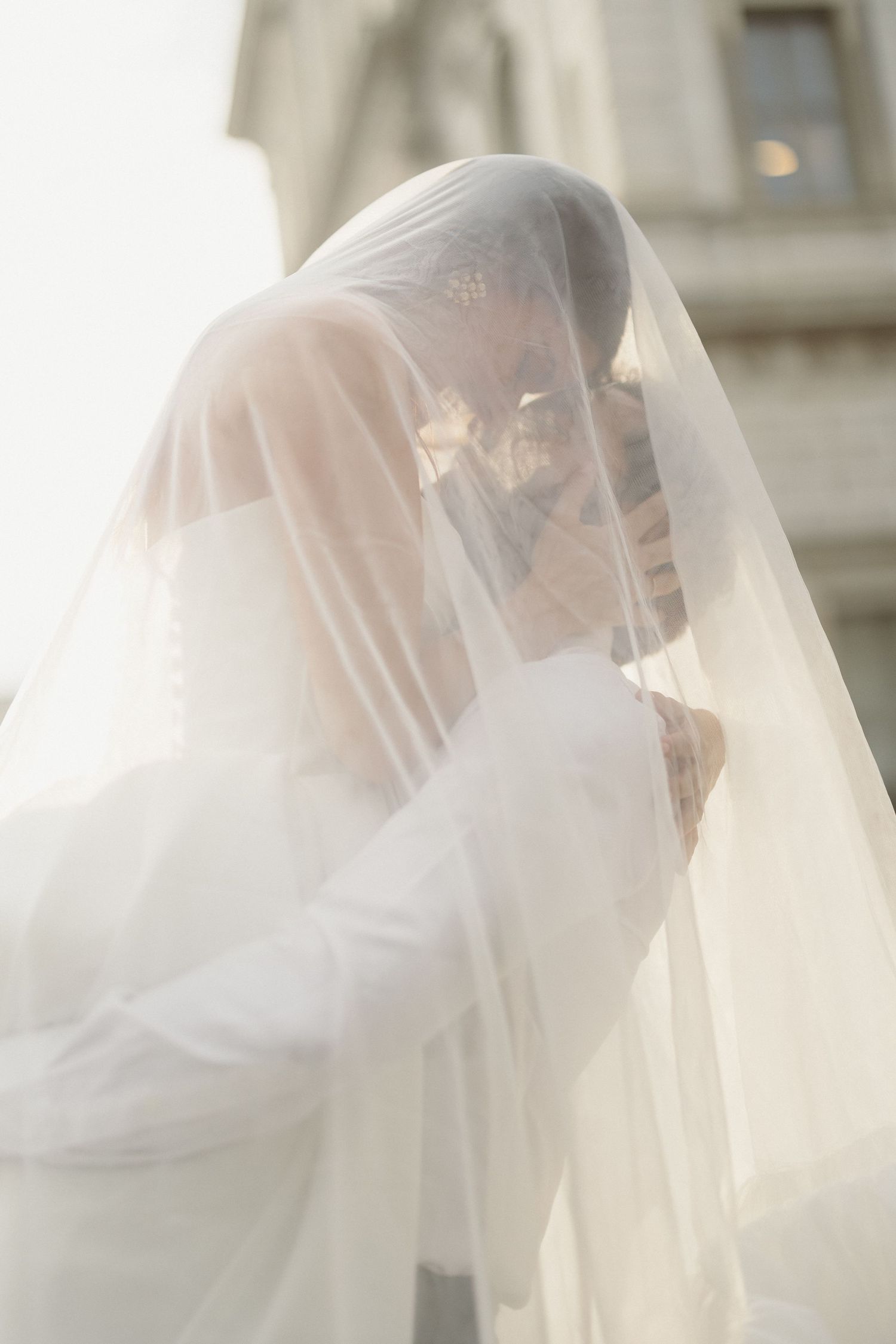 Romantic bride and groom kissing beneath flowing white veil in soft natural light.