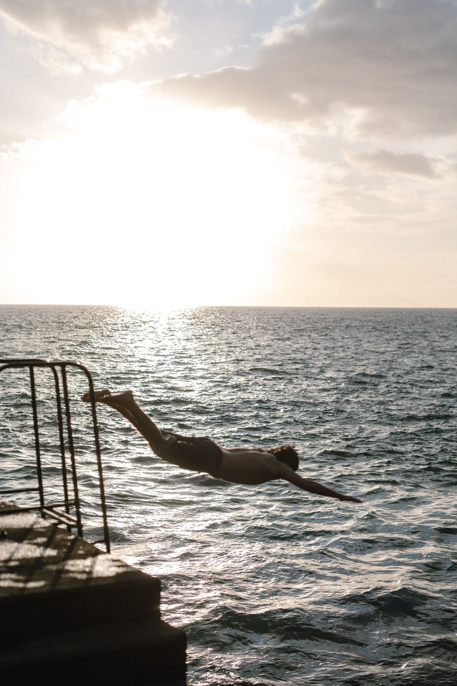 Person diving off a pier into the ocean at sunset, silhouetted against glimmering water and cloudy sky.