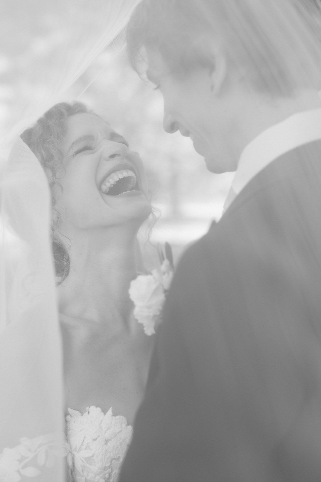 Black and white photo of a laughing bride and groom sharing an intimate moment under a wedding veil.