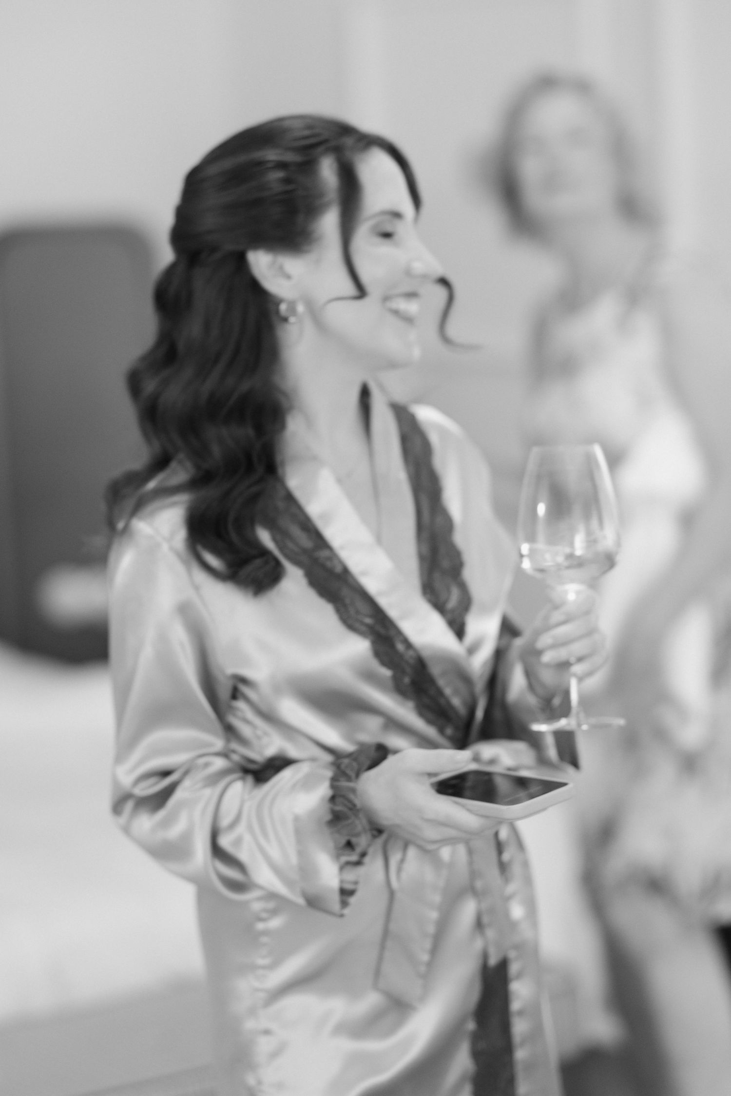 Black and white photo of a smiling woman in a satin robe holding a wine glass during bridal preparations.