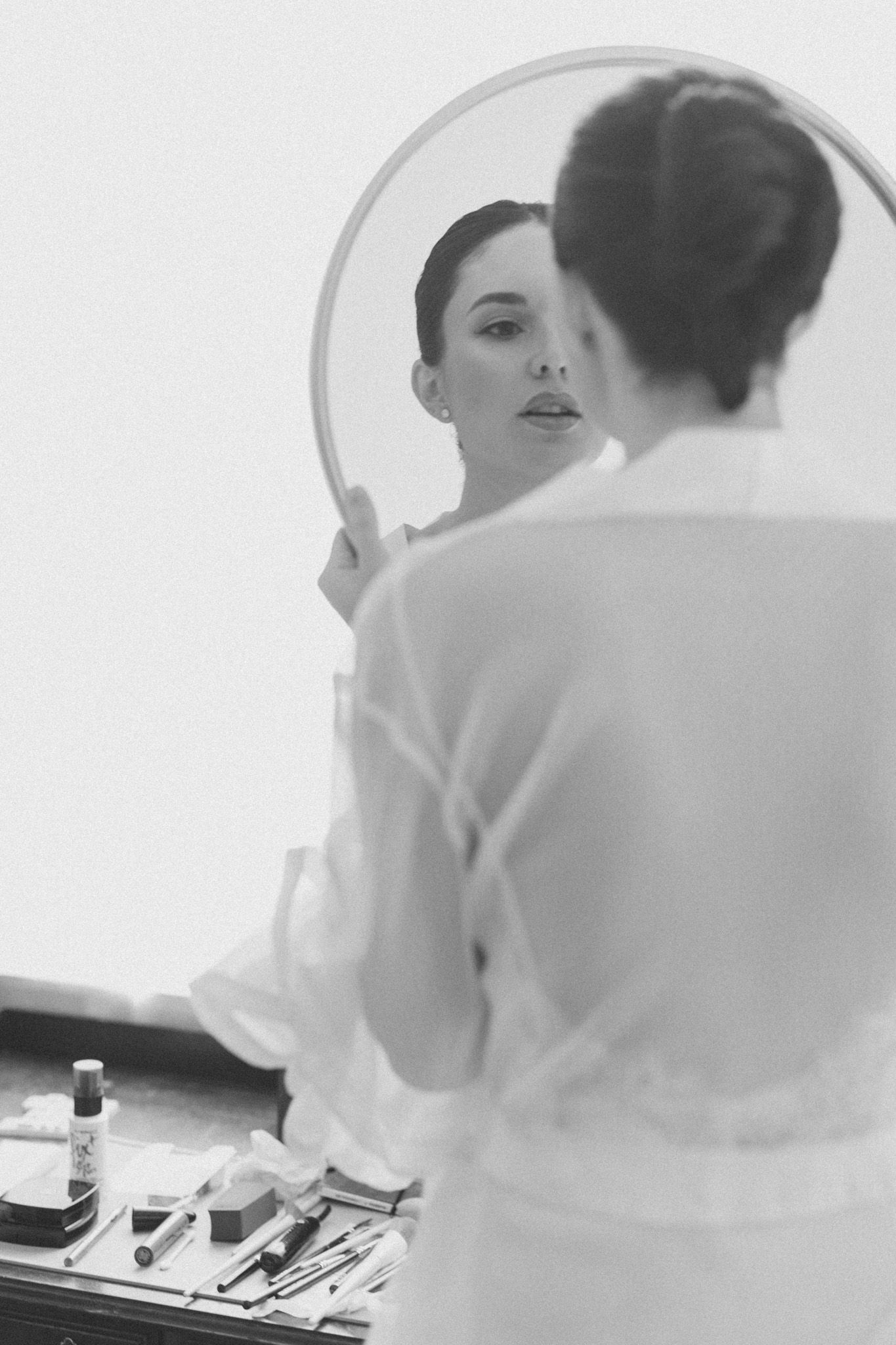Woman in white robe applying makeup while looking at her reflection in a round mirror, black and white photo.