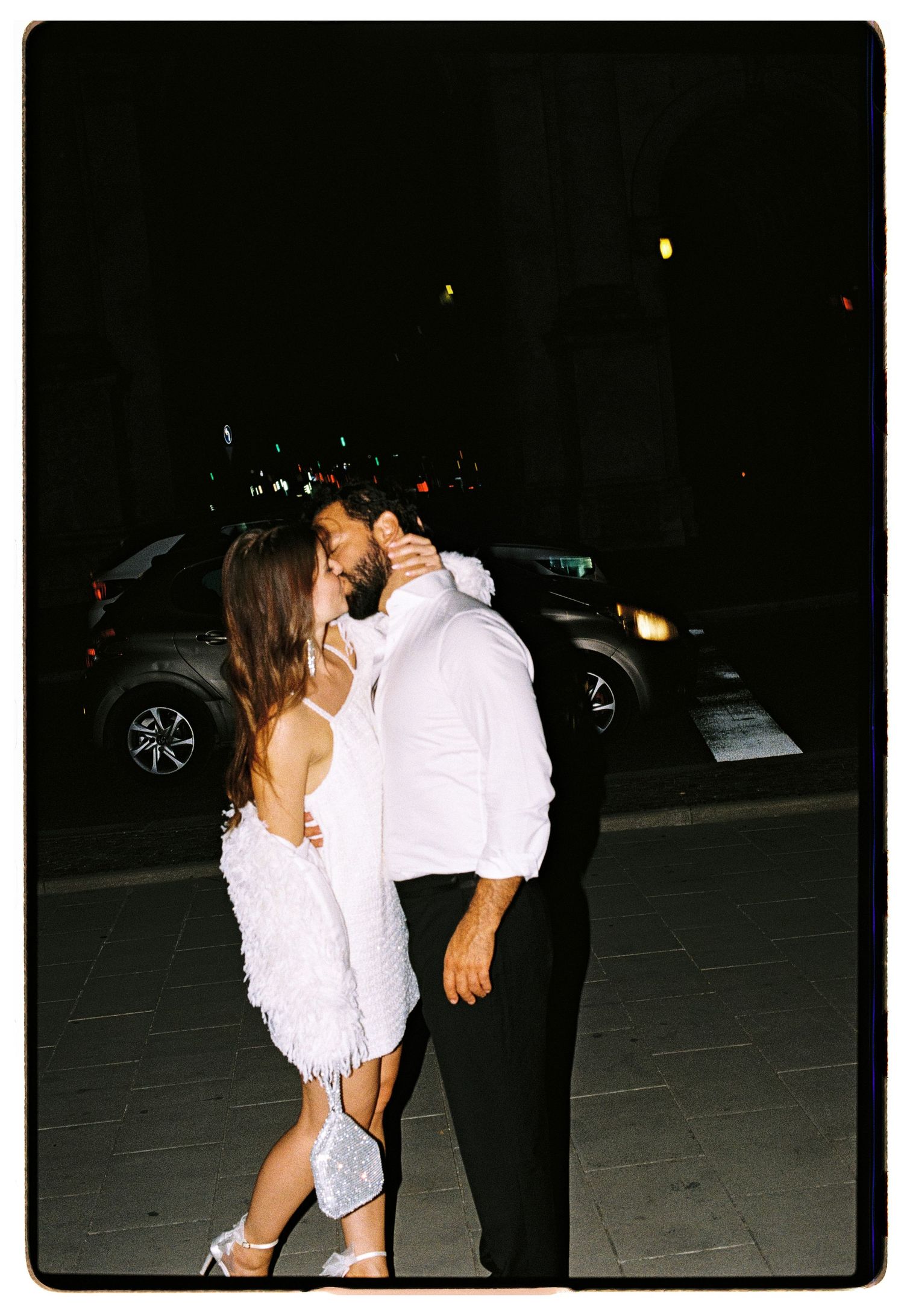 Romantic couple sharing a kiss outdoors at night, woman in white feathered dress and man in white shirt and black pants.