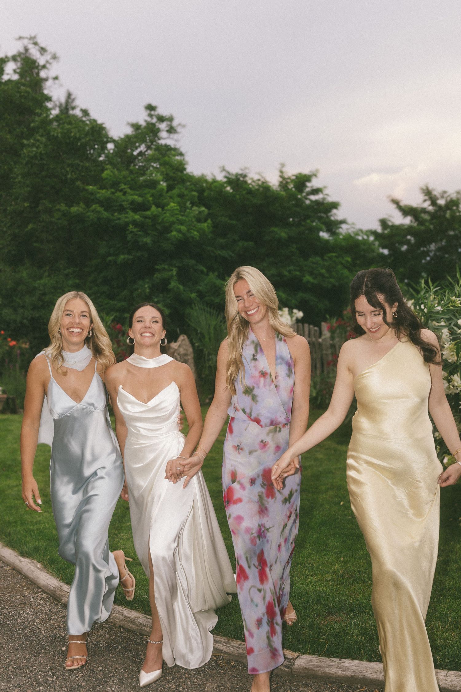Four women in elegant satin gowns walk together outdoors at a wedding celebration.