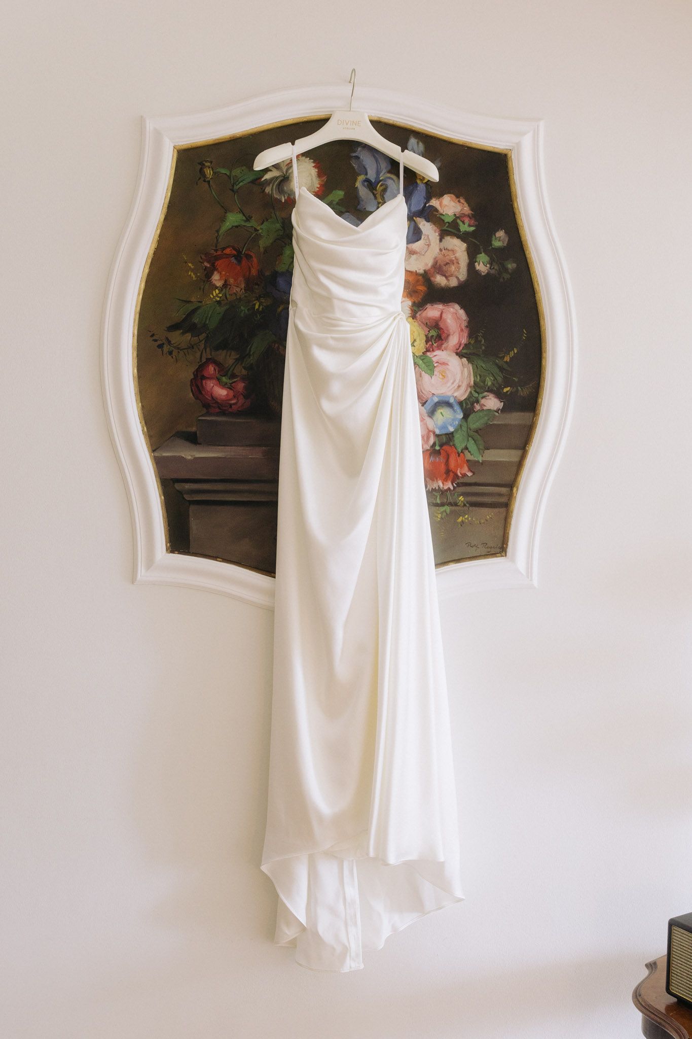 Elegant white bridal gown hanging on a hanger against a floral painting in a white ornate frame.