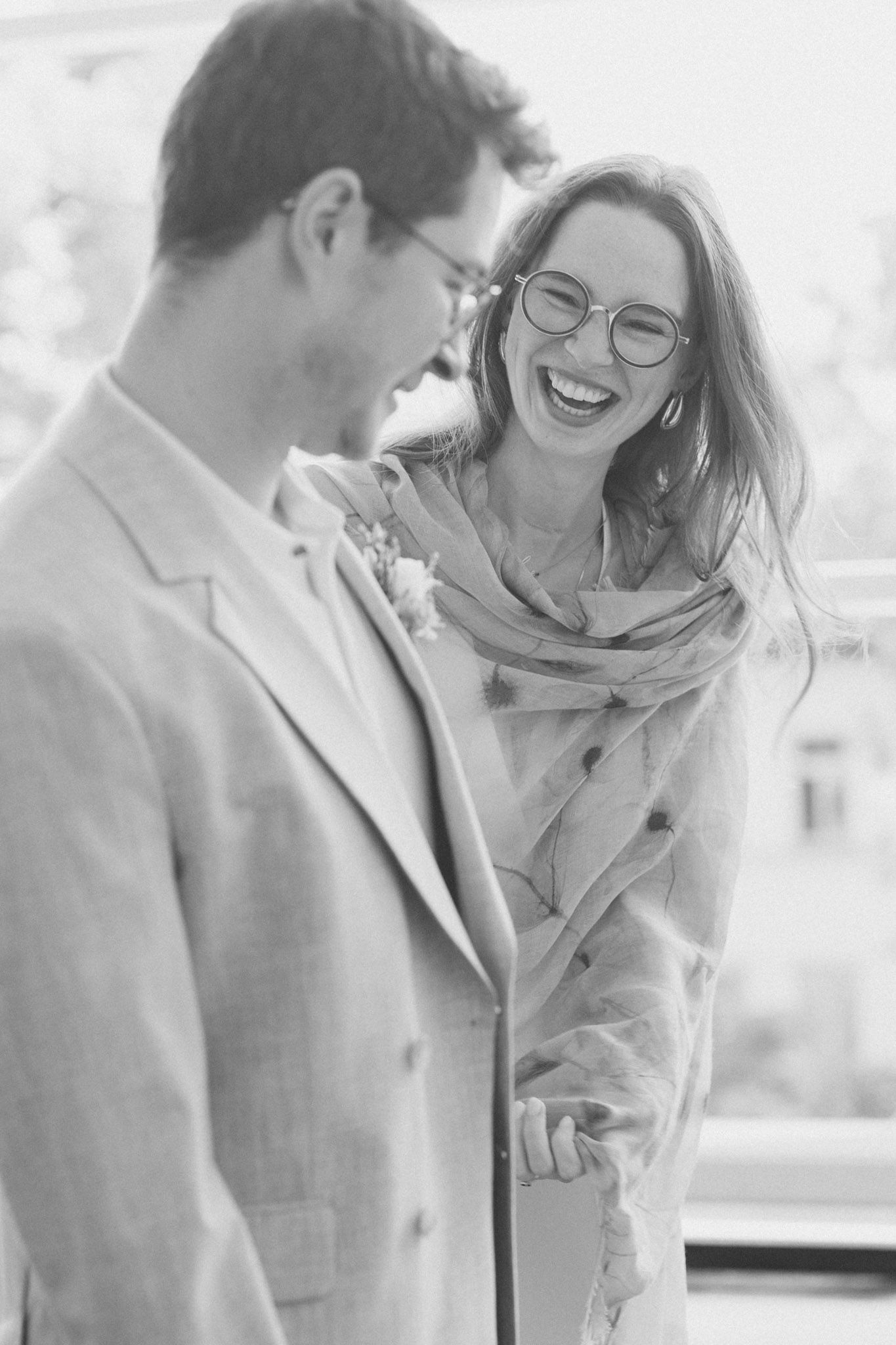 Black and white photo of a laughing bride in glasses and groom in suit sharing a joyful moment together.