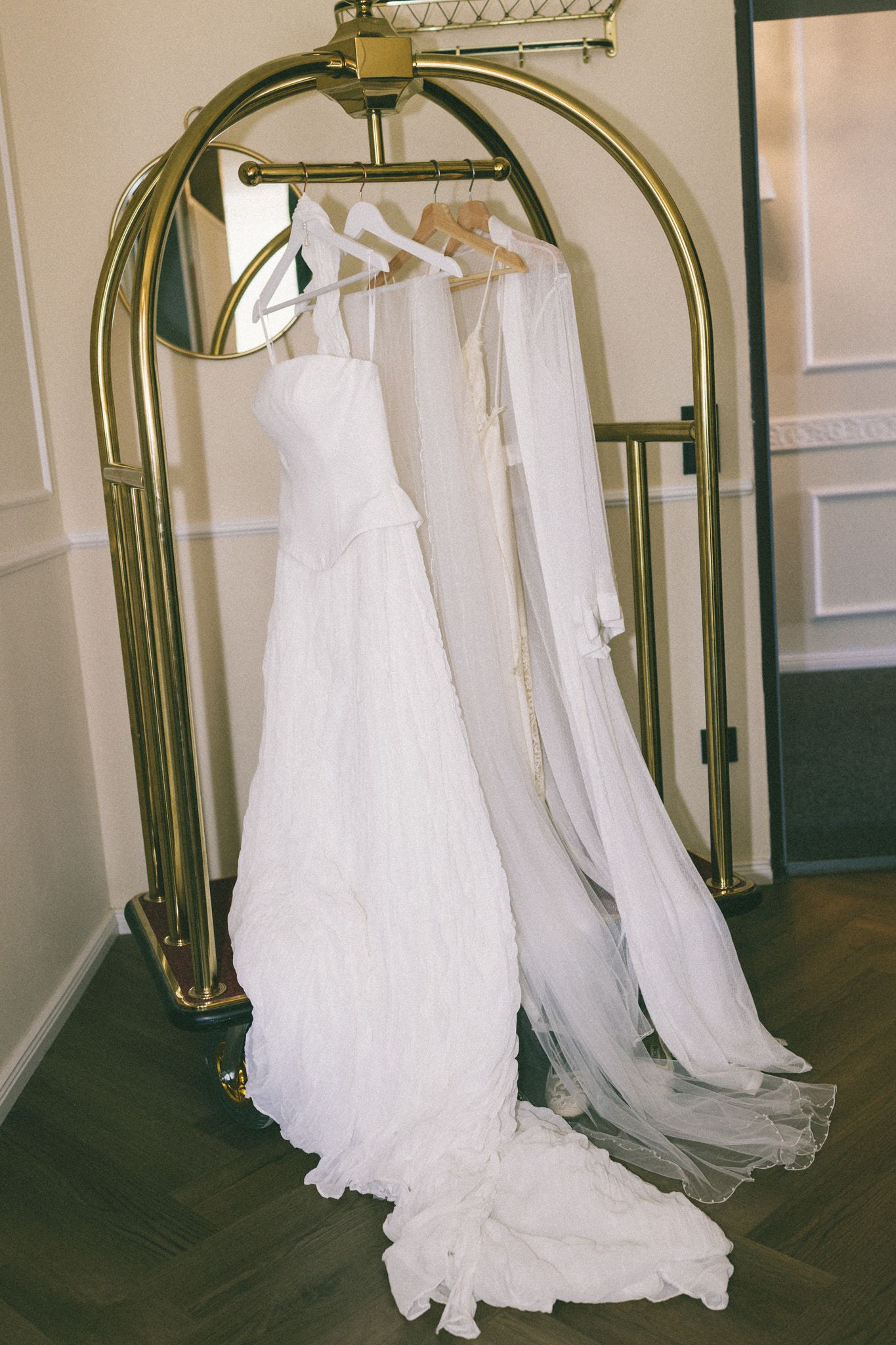 White wedding dress and veil hanging on a gold luggage rack in an elegant hotel room.