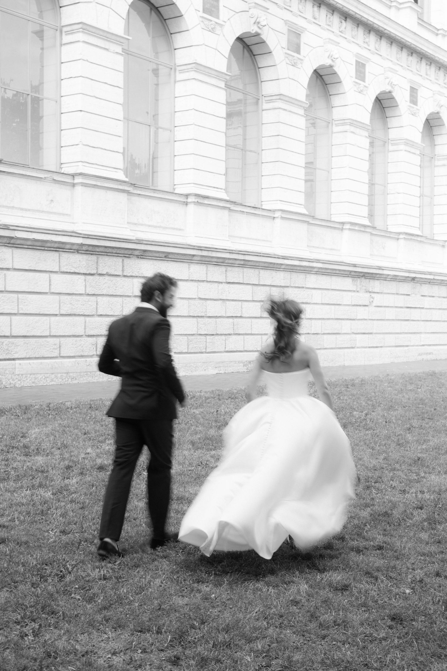 Black and white photo of bride and groom running across grass beside grand building.