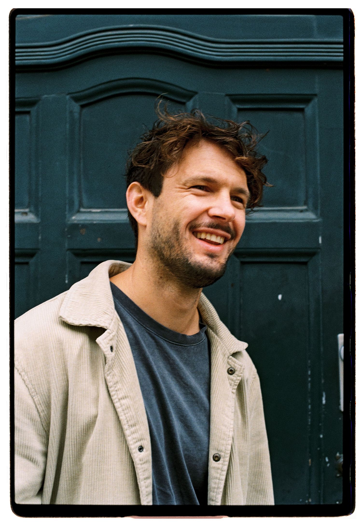 Smiling man with curly hair and beard standing in front of a teal door wearing a beige jacket.