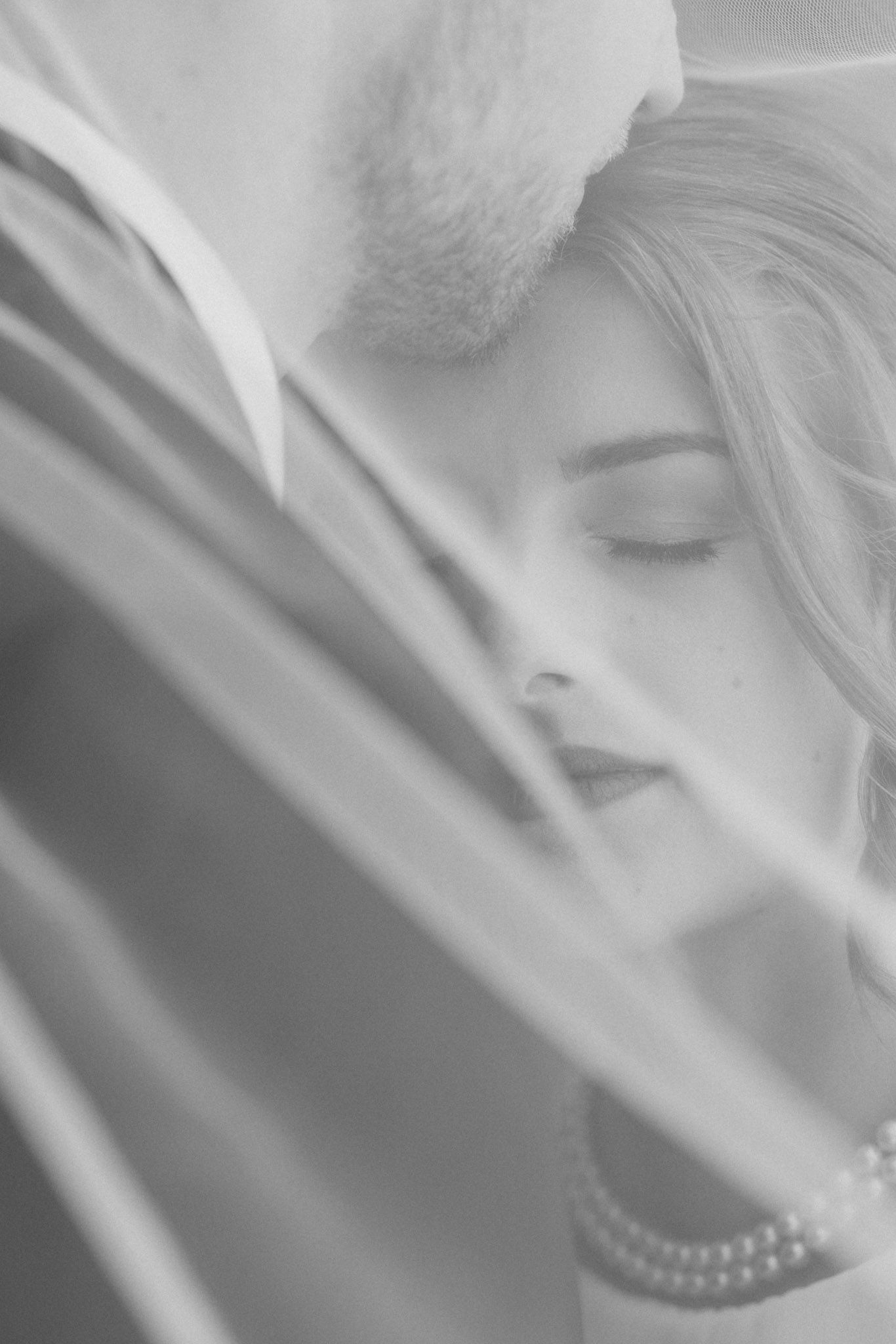 Black and white wedding photo of bride and groom sharing an intimate moment under a veil, bride wearing pearl necklace.