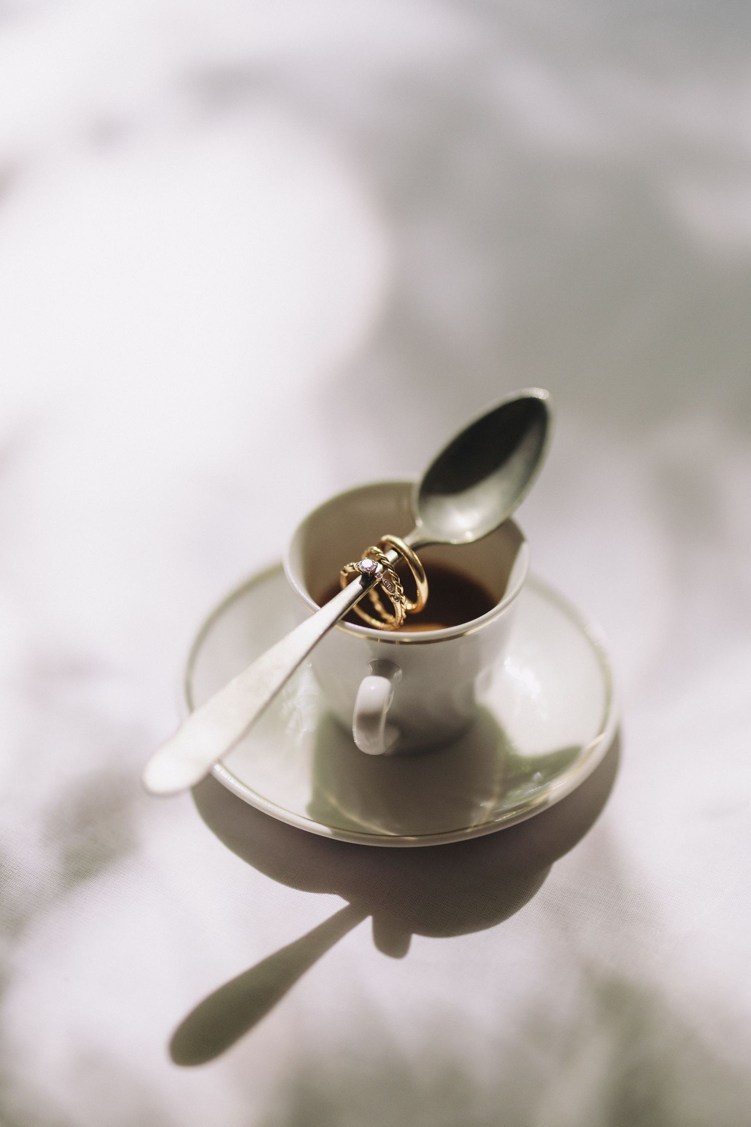 Gold wedding rings resting in a white espresso cup with spoon on a saucer in soft natural light.