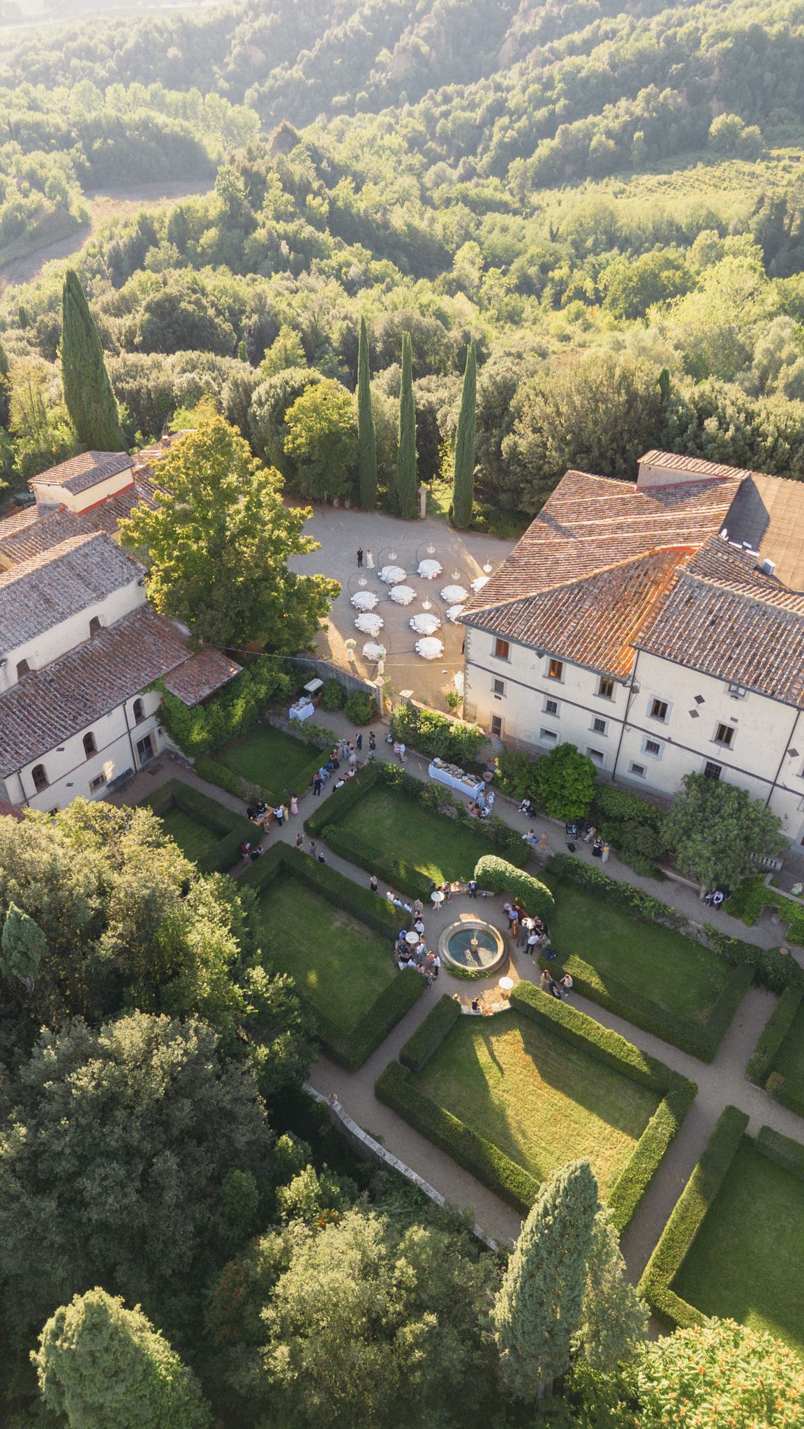 Aerial view of a stunning Italian villa with manicured gardens, fountain, and terracotta rooftops surrounded by lush Tuscan hills.