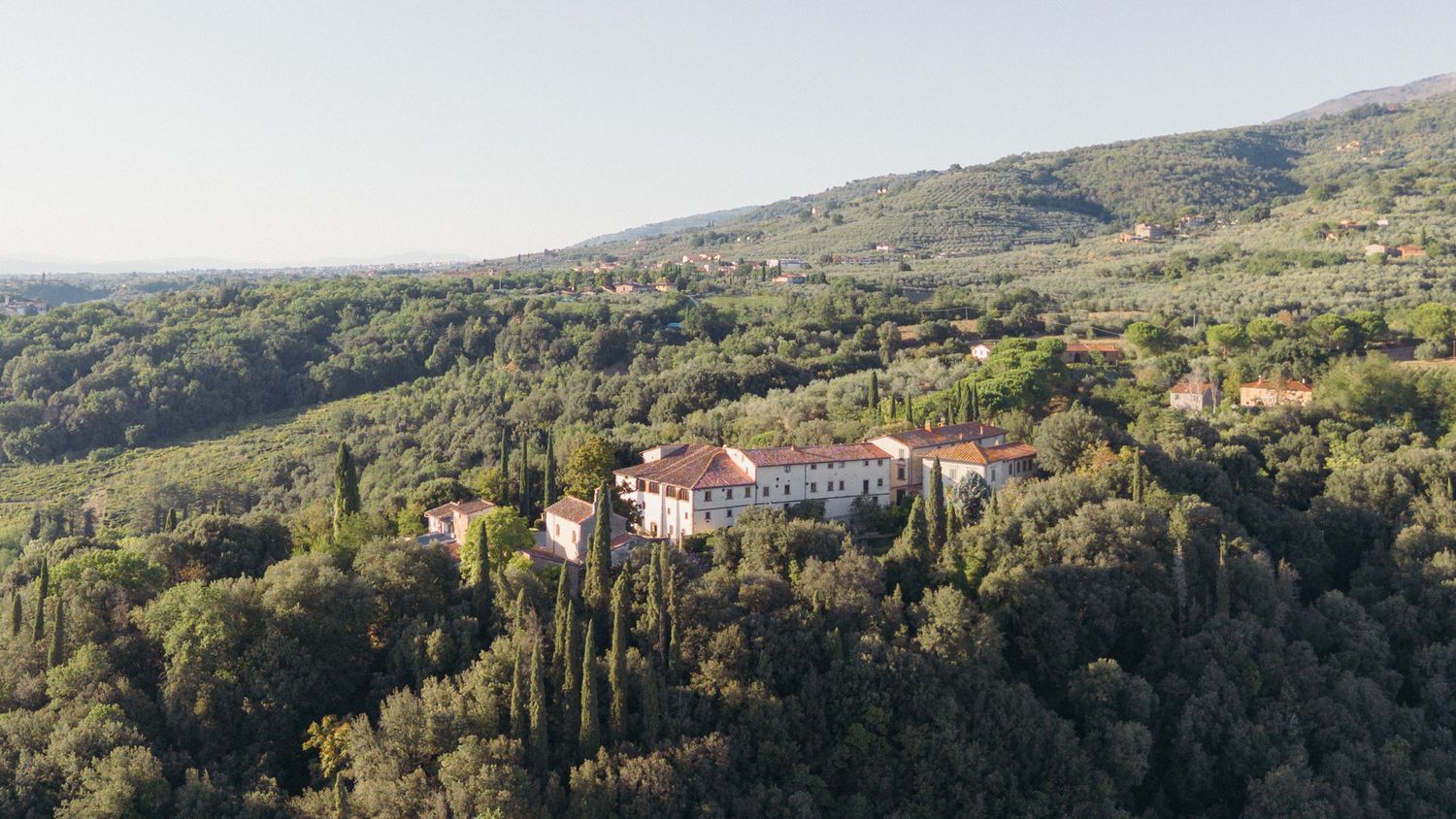 Aerial view of a historic Italian villa surrounded by lush green forests and cypress trees on a hillside.