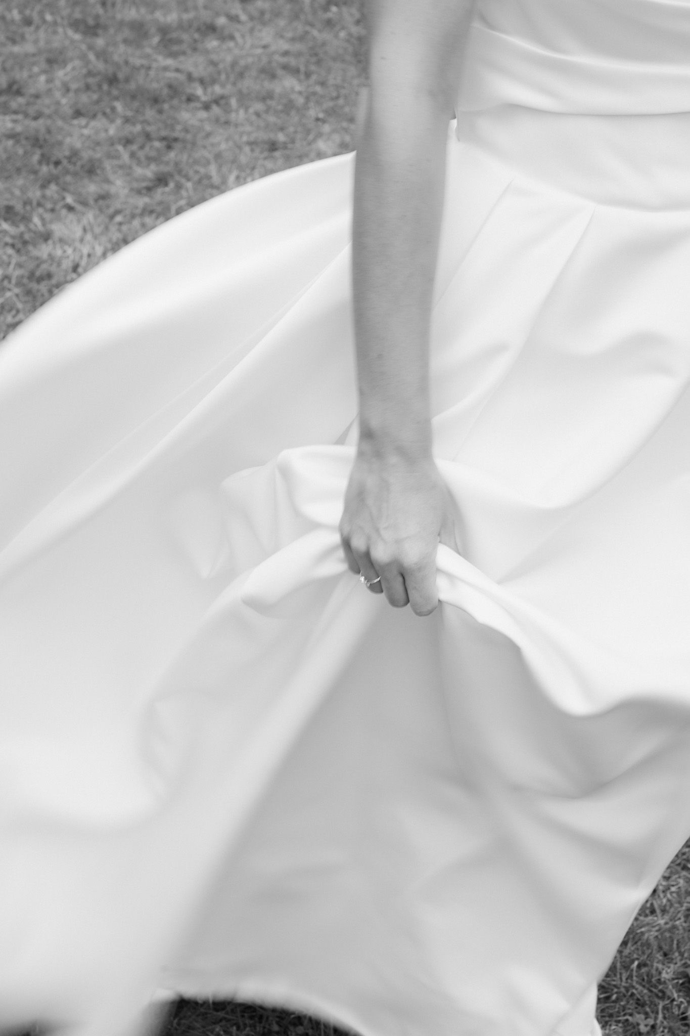 Black and white photo of a bride holding her flowing white wedding dress skirt outdoors on grass.