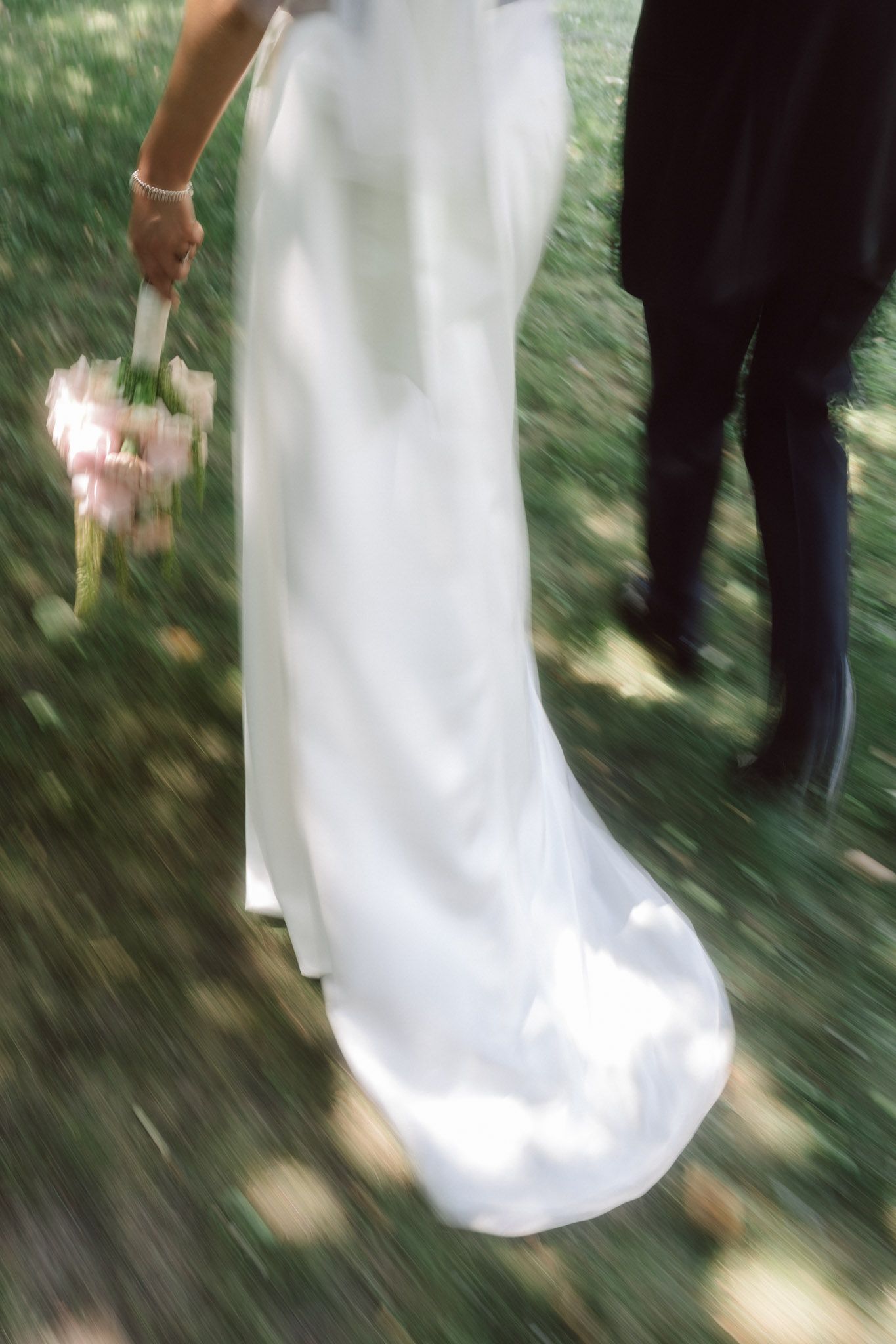 Bride in white gown holding pink bouquet walks with groom in black suit on green grass, motion blur effect.