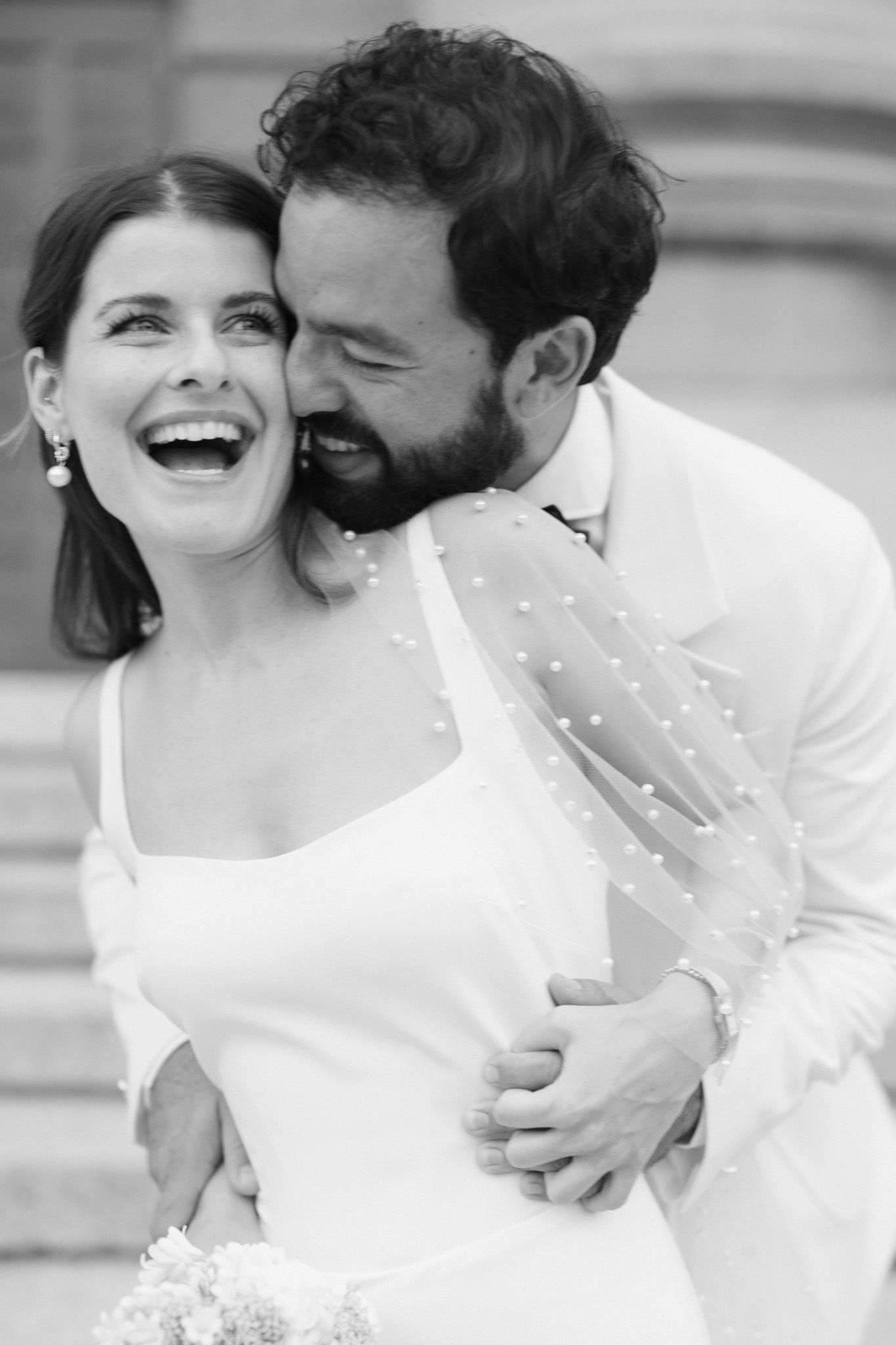 Black and white wedding photo of laughing bride in pearl veil embraced by bearded groom in white suit.
