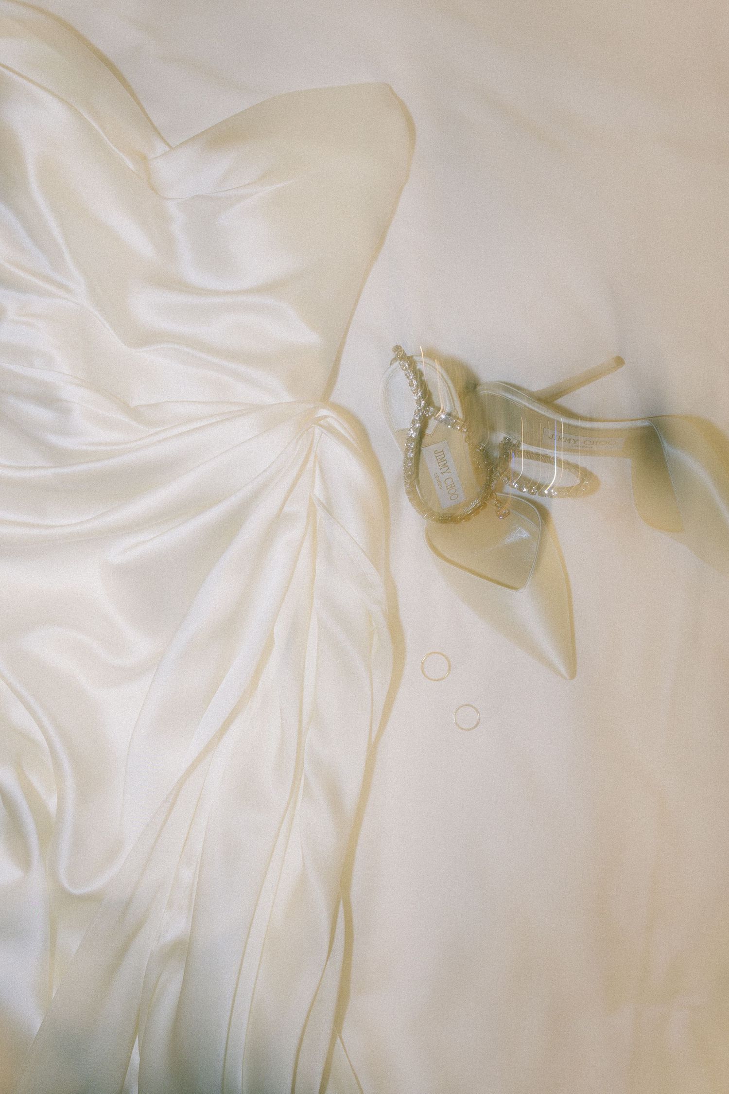 Close-up of a white satin wedding dress laid flat with crystal-embellished bridal heels and delicate pearl jewelry.