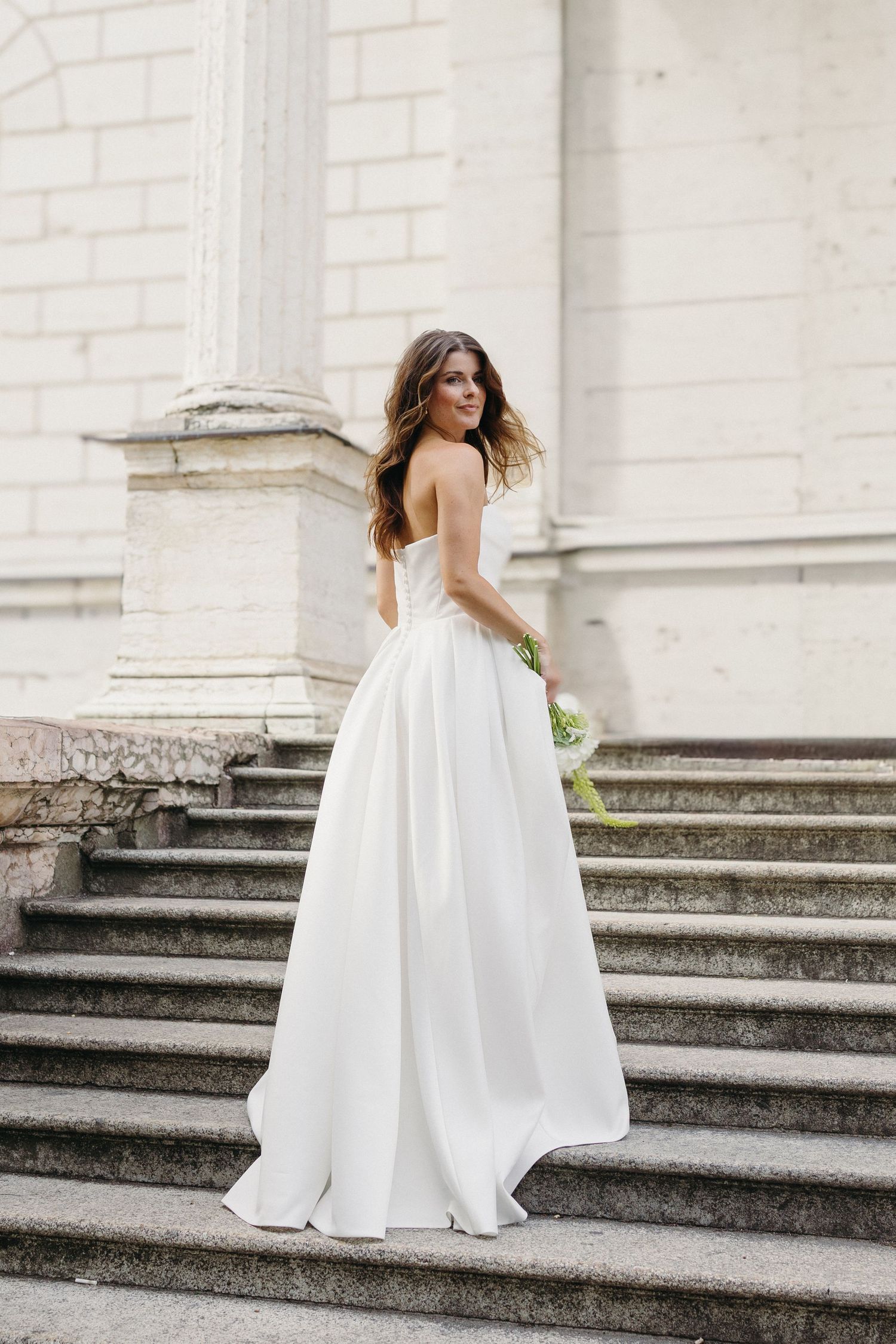 Bride in strapless white ball gown holding bouquet on stone steps of historic building.