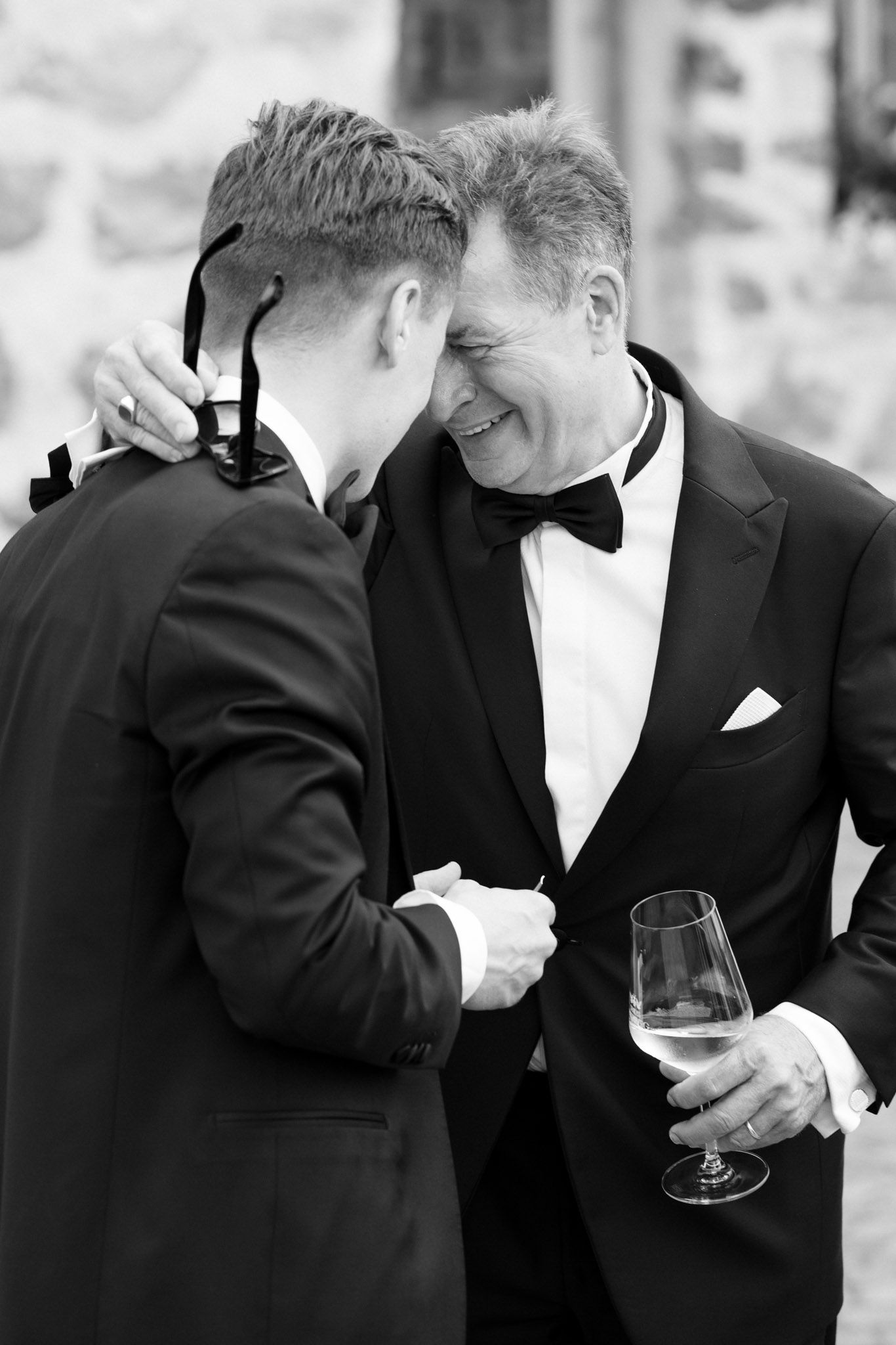 Two men in tuxedos share a joyful moment, one holding a wine glass, in a black and white photo.