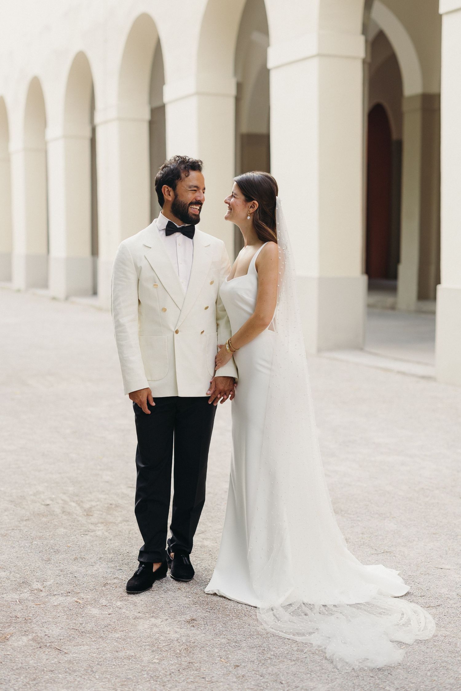 Smiling bride and groom in white tuxedo holding hands beneath arched courtyard arcade.