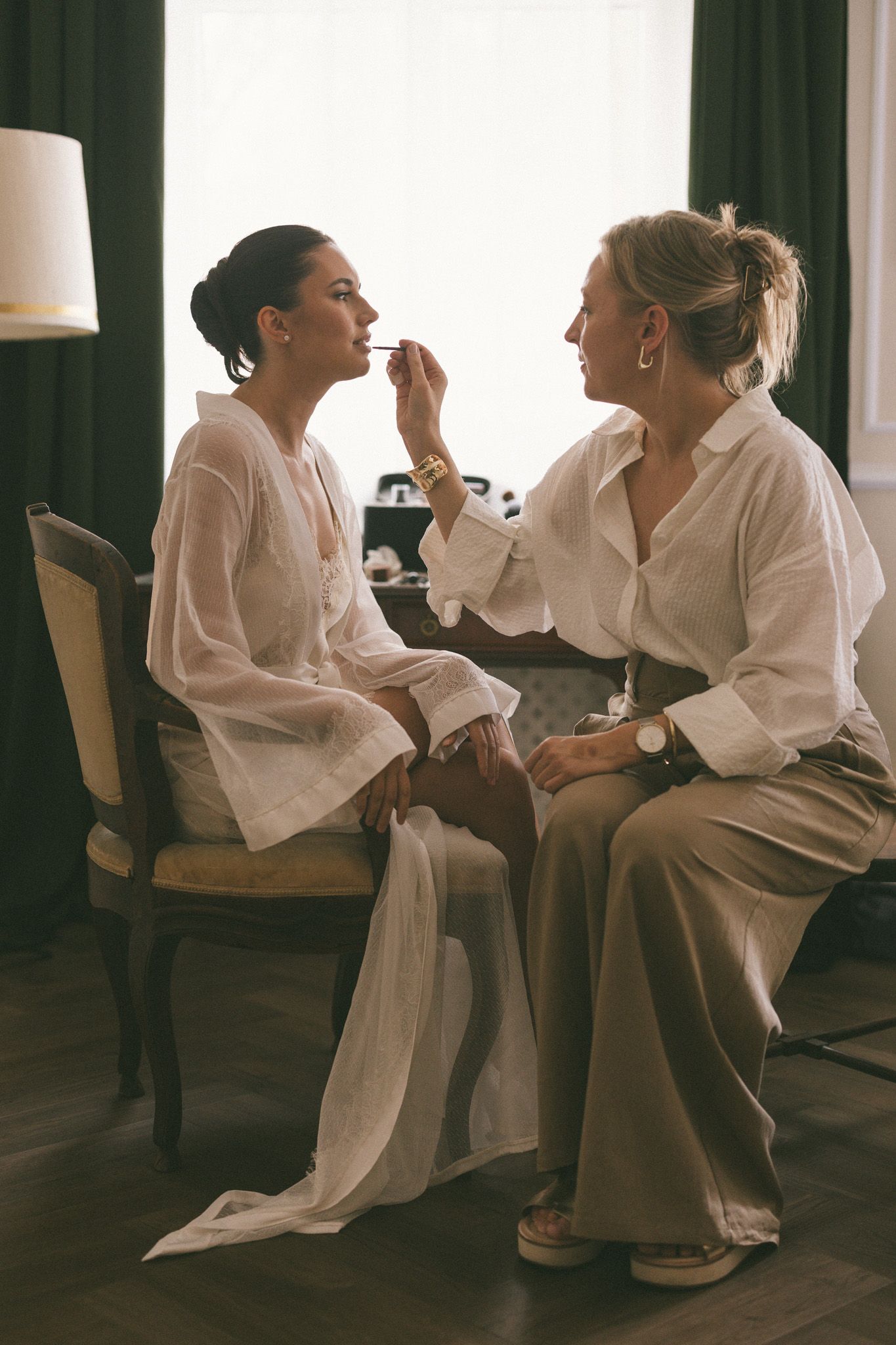 Two women in white blouses sit together as one applies lipstick to the other in an elegant room.