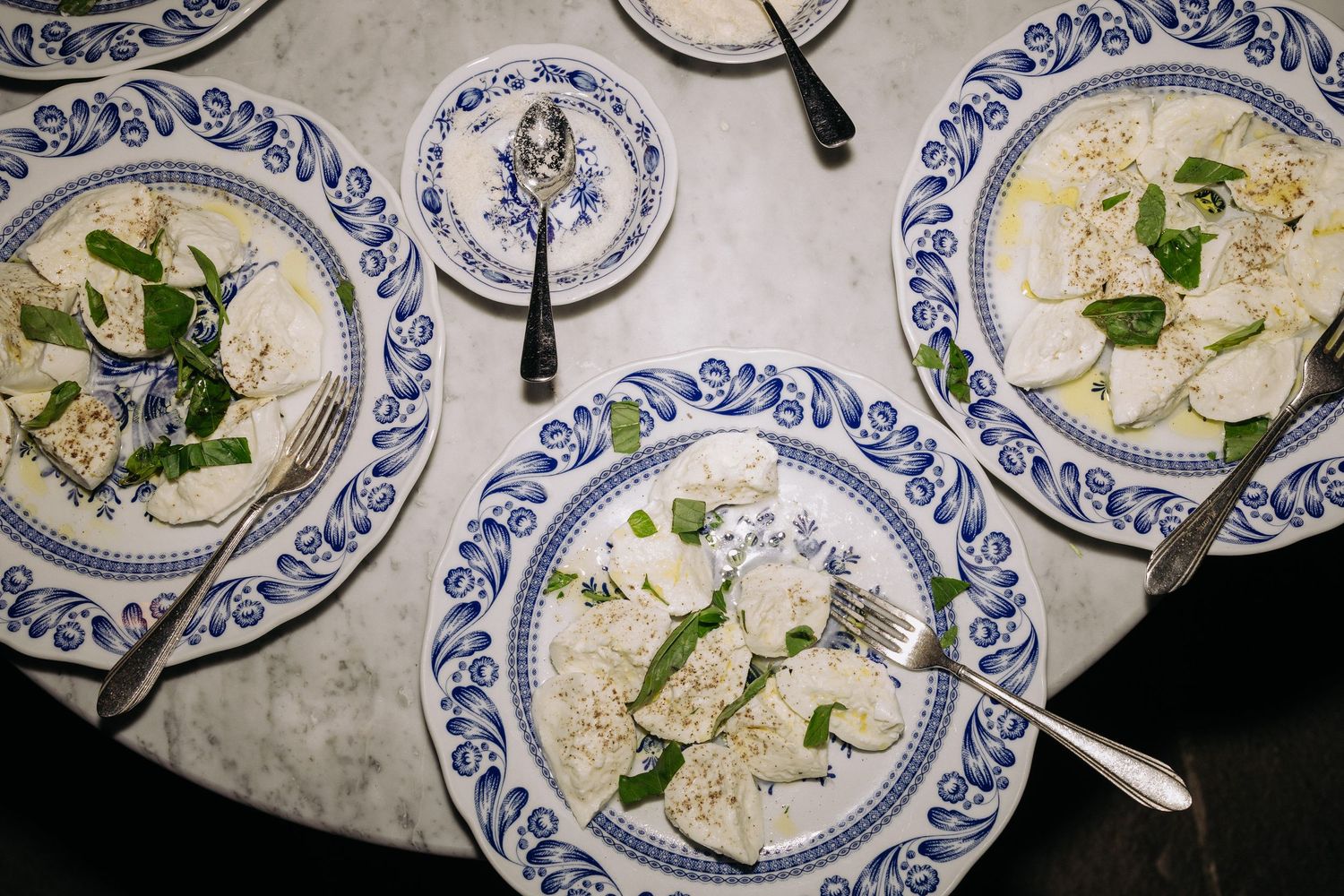 Overhead view of elegant blue and white plates with caprese salad topped with fresh basil and mozzarella on a marble table.