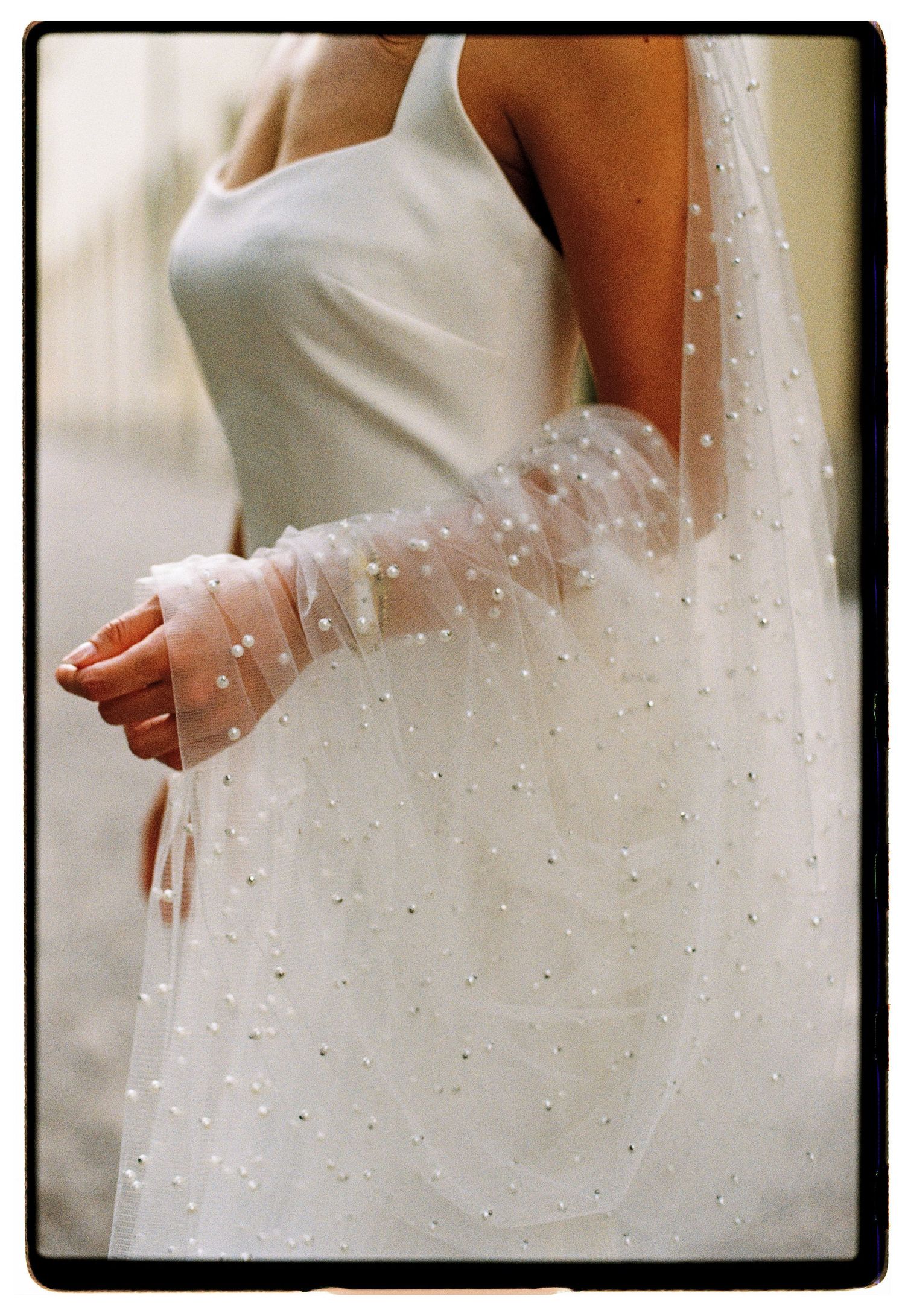 Detail shot of bride in white satin wedding dress with pearl-embellished tulle veil draped over her arm.