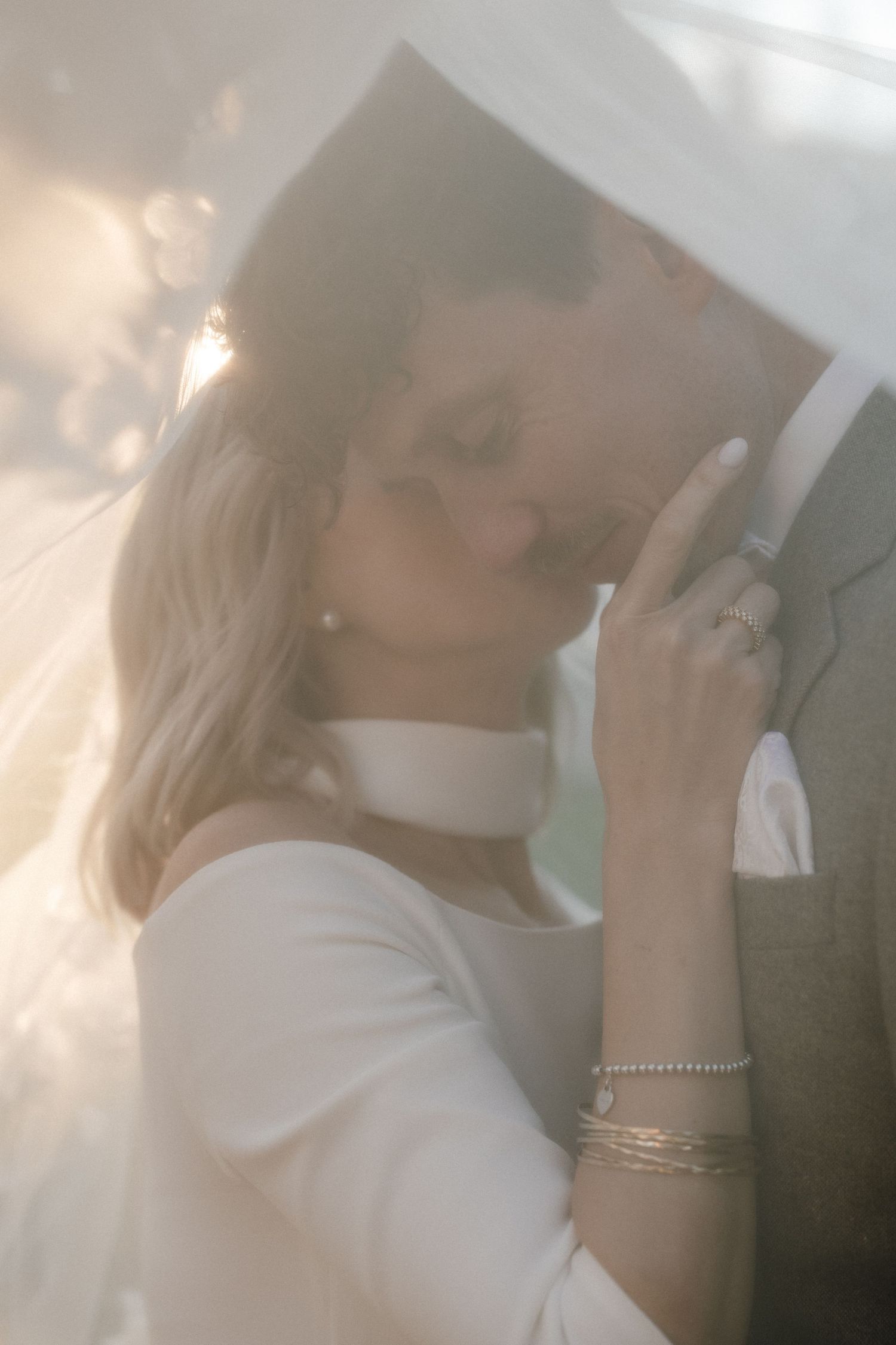 Romantic wedding couple sharing an intimate moment under a veil, bride in white dress with bracelets and ring.