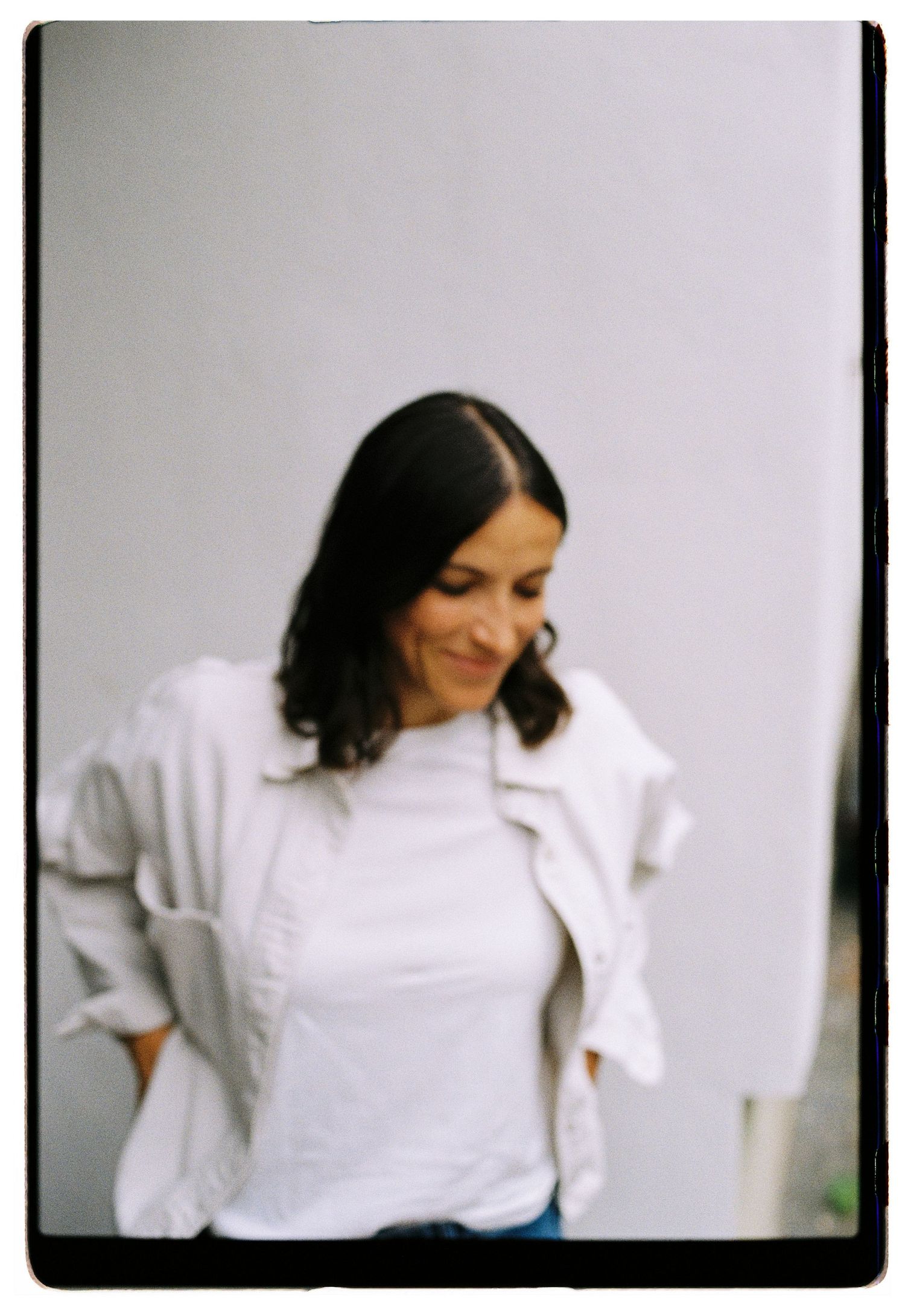 Stylish woman in white t-shirt and jacket smiling downward against a light gray background.