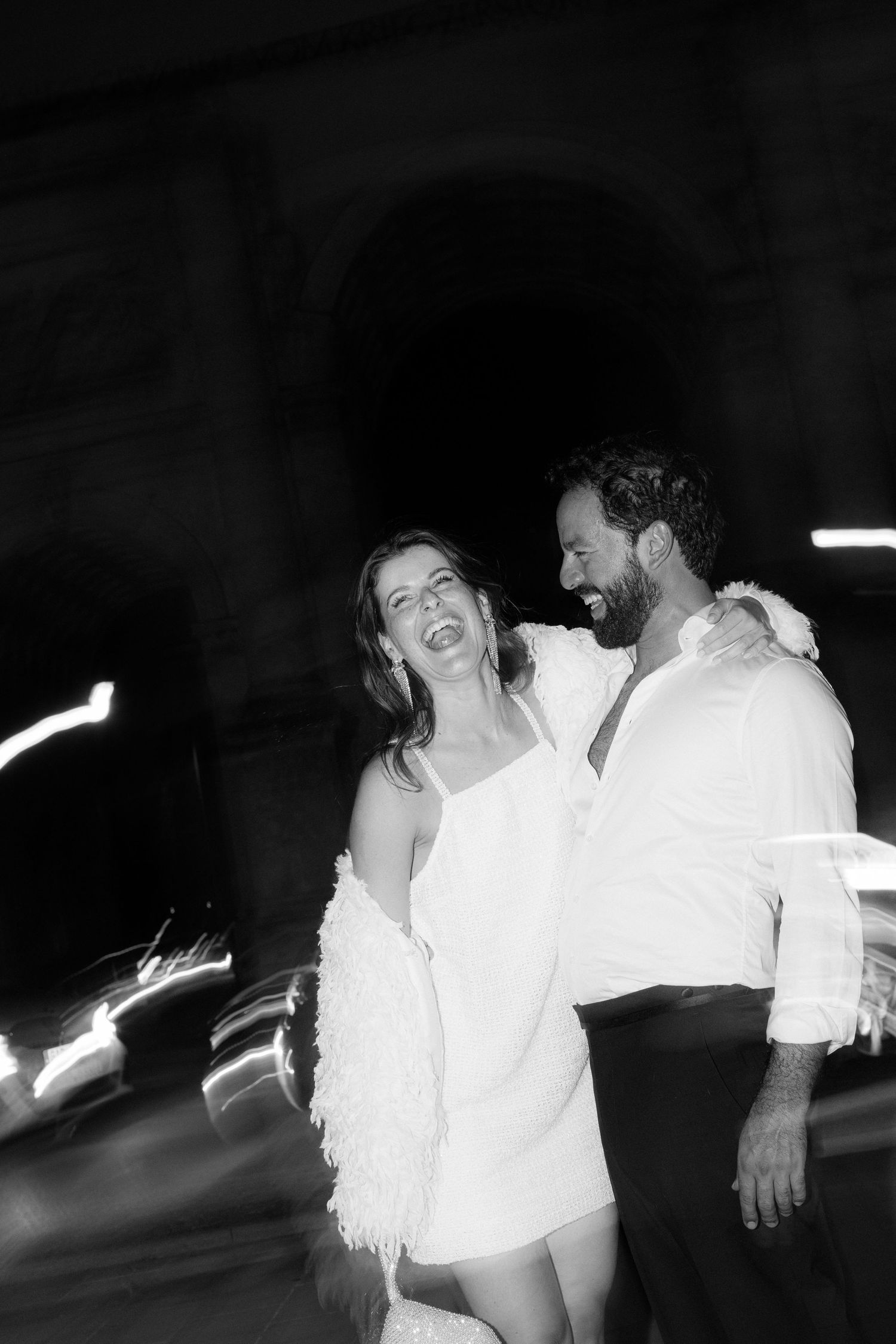 Laughing bride in white mini dress with groom in white shirt at night with light streaks, black and white.