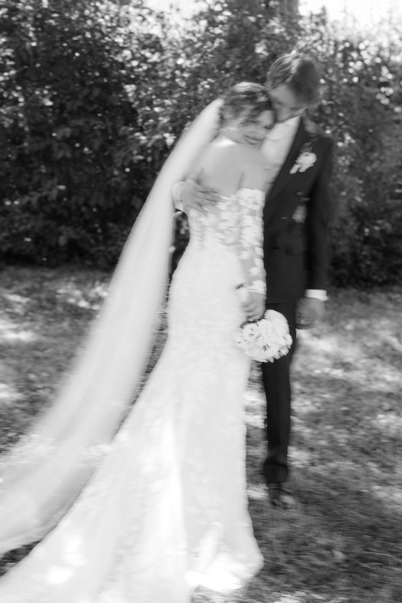 Black and white photo of a bride and groom sharing a tender moment outdoors, bride holding bouquet in lace gown with veil.