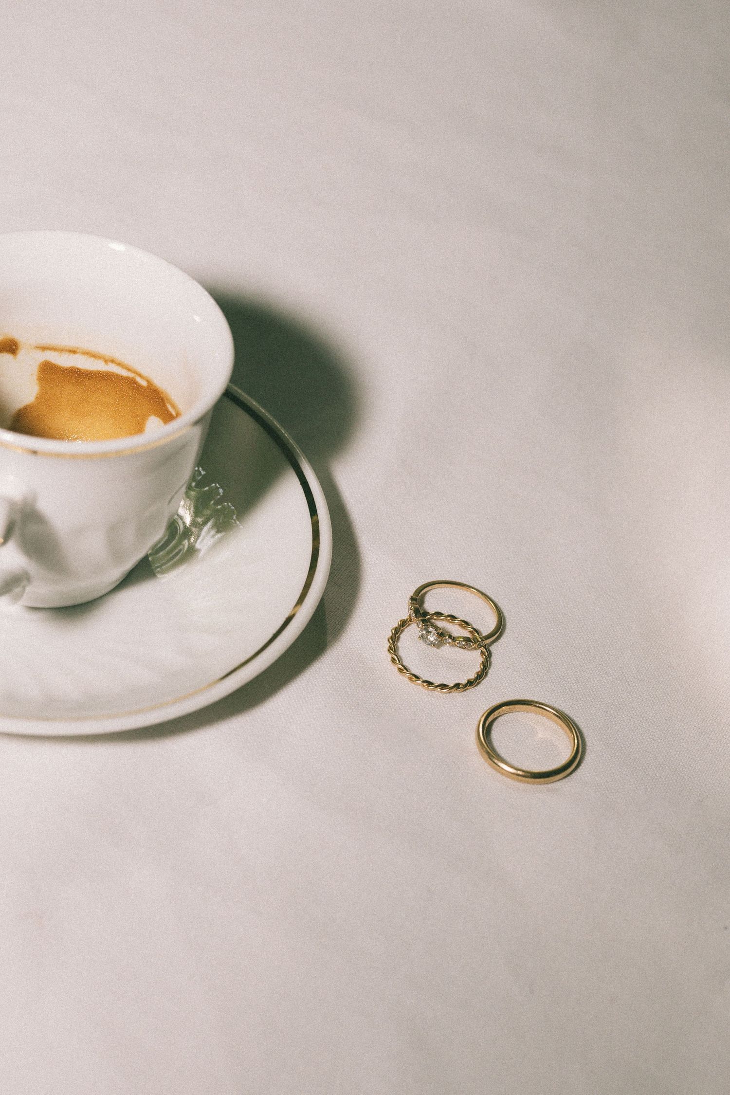 Gold wedding bands beside a white espresso cup on saucer with coffee on a light neutral surface.