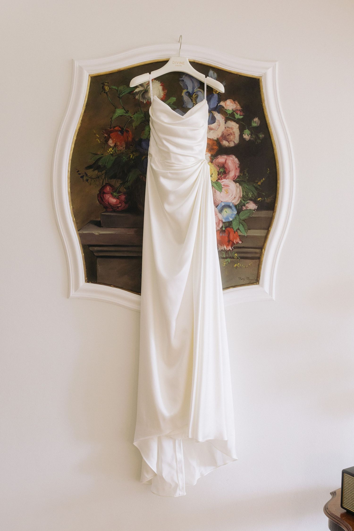 Elegant ivory satin wedding gown hanging on a white hanger in front of a vintage floral painting on a wall.