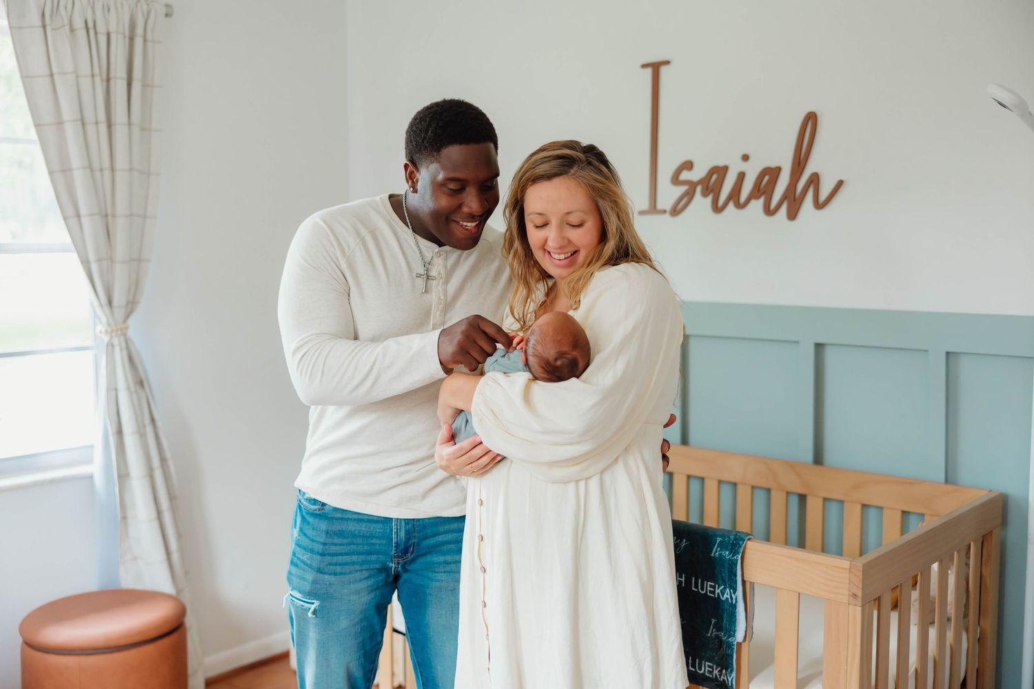 Smiling couple holding newborn baby in nursery with Isaiah name sign and wooden crib.