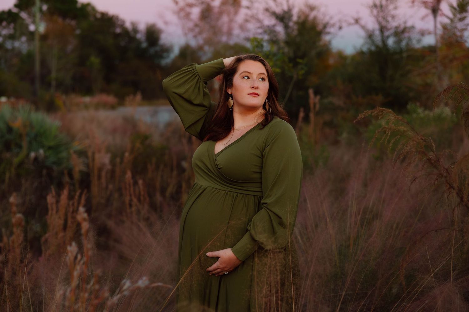 Pregnant woman in green wrap dress posing in golden hour light among tall grasses.